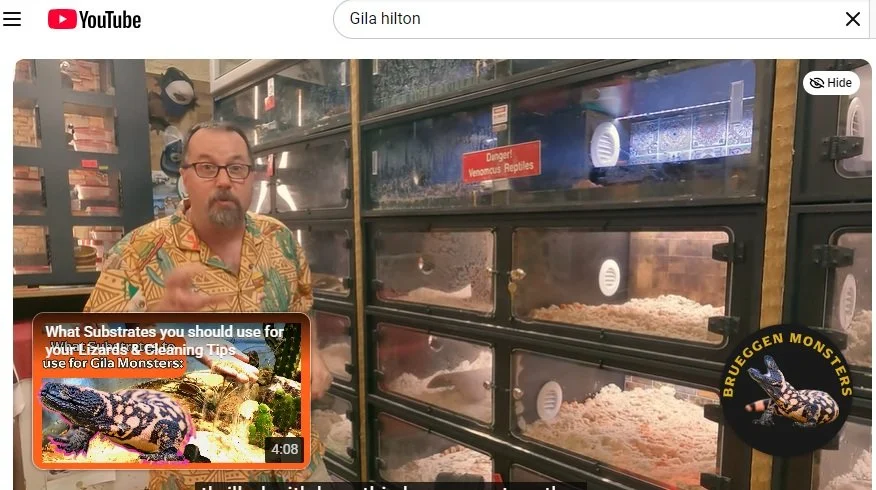 A man with glasses and a patterned shirt standing in front of multiple reptile enclosures, with a sign warning about venomous reptiles, and a logo for 'Brueggen Monsters' featuring a Gila Monster.