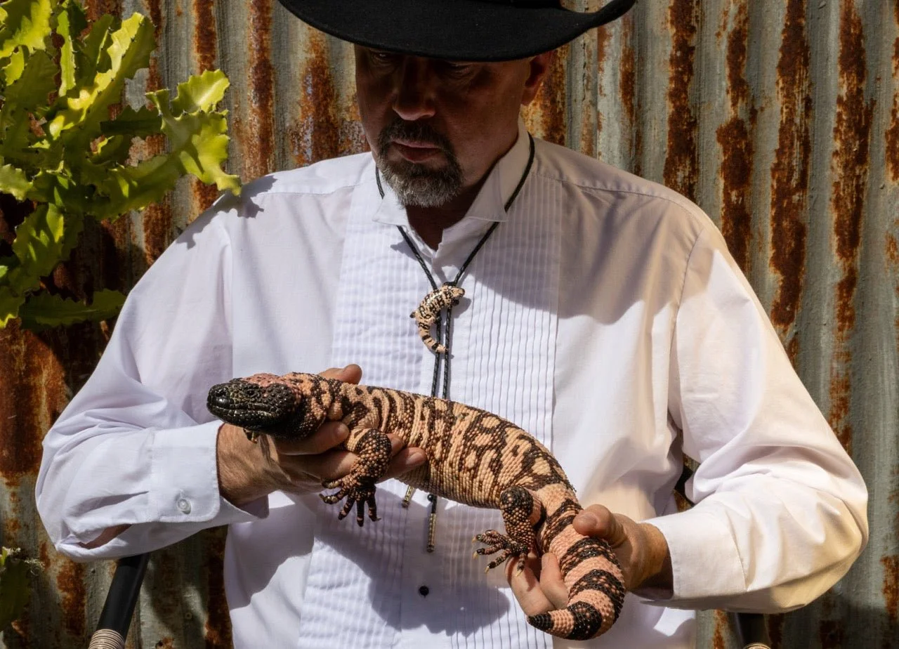 A man wearing a white shirt and black hat holding a colorful lizard with black and pinkish markings, standing in front of a rusty metal wall and some green plants.