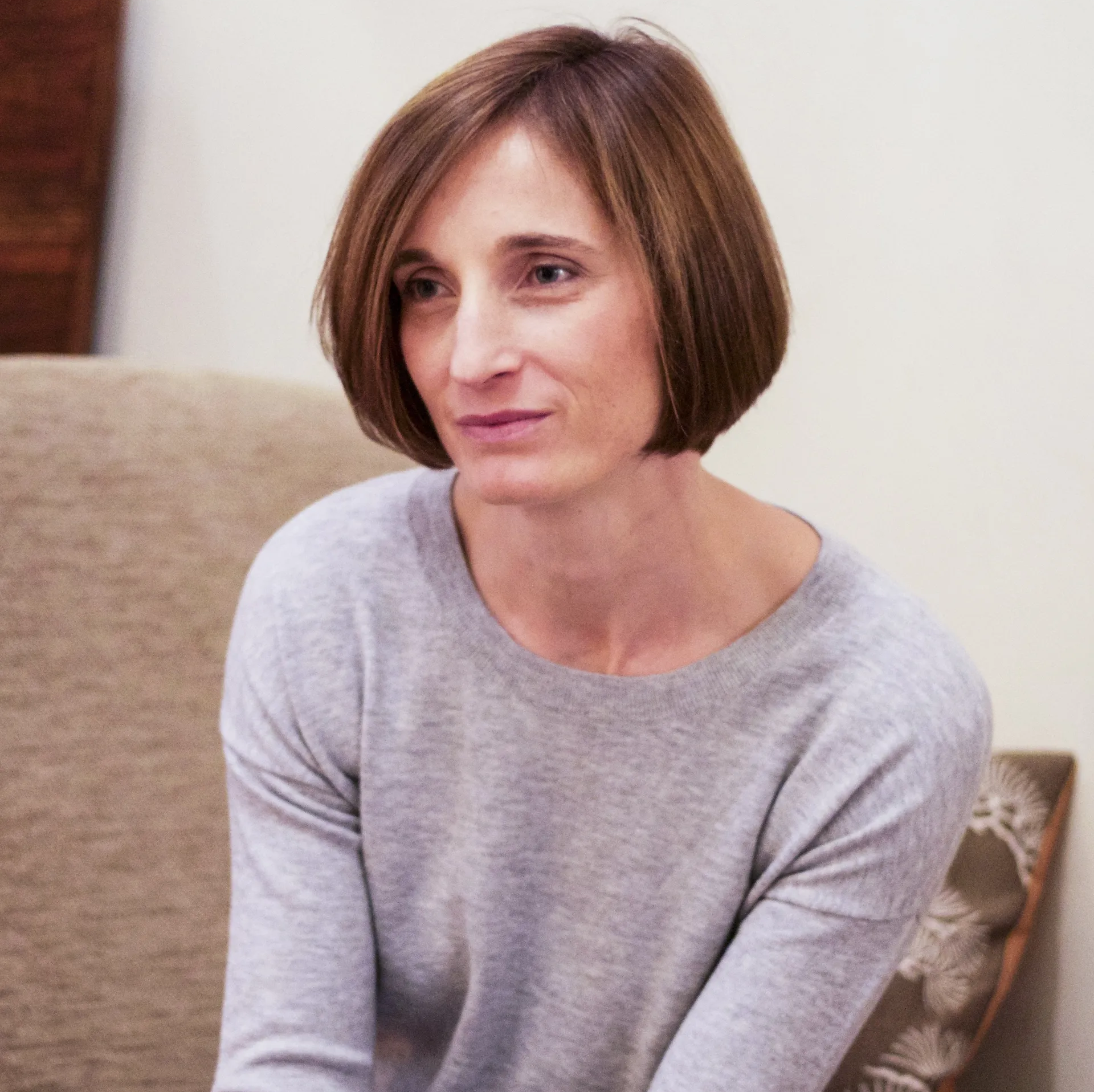 A woman with short, light brown hair, wearing a light grey sweater, sitting on a beige couch in a living room with wooden furniture and a rug.