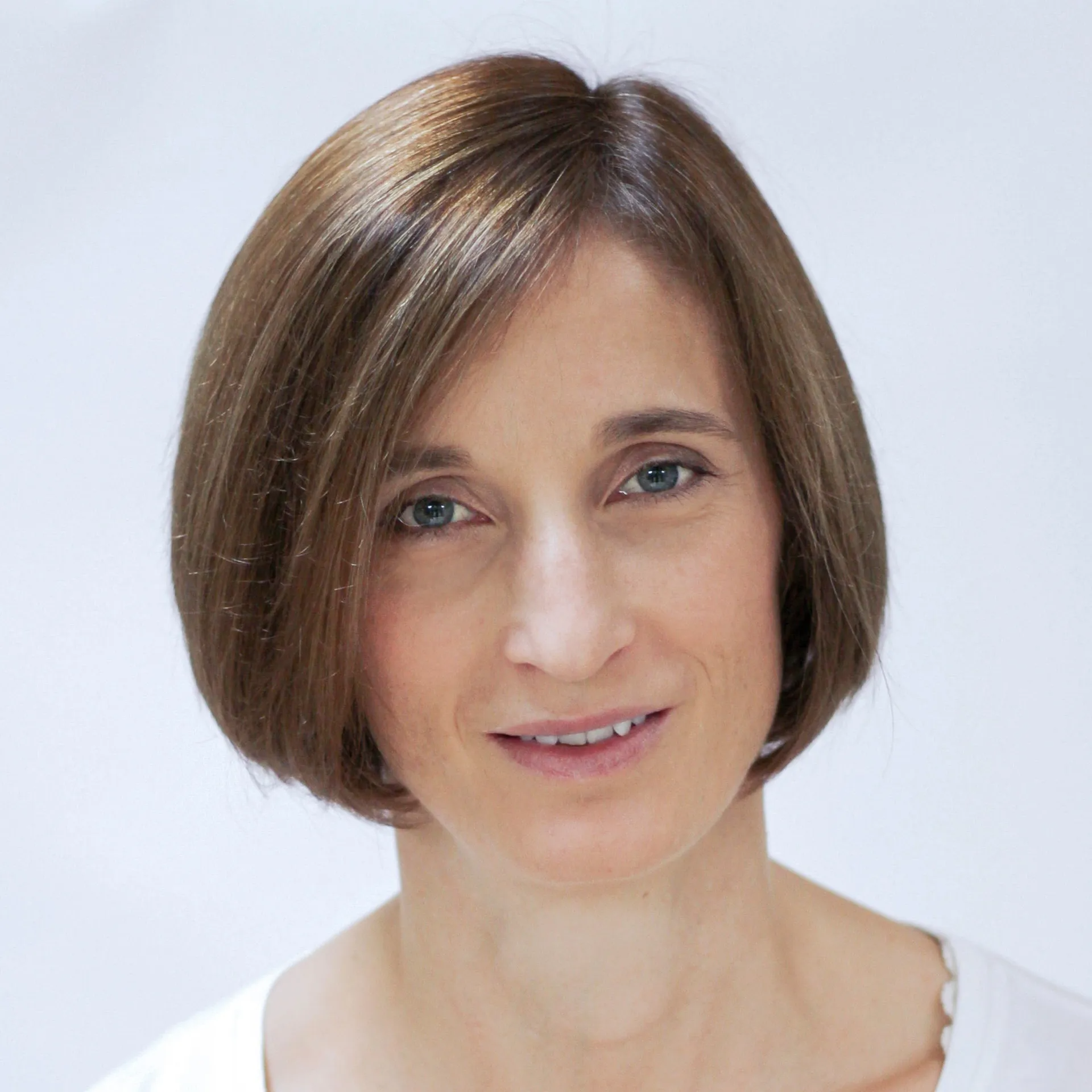 A woman with short, light brown hair, fair skin, and blue eyes is smiling slightly at the camera. She is wearing a white top. The background is plain white.