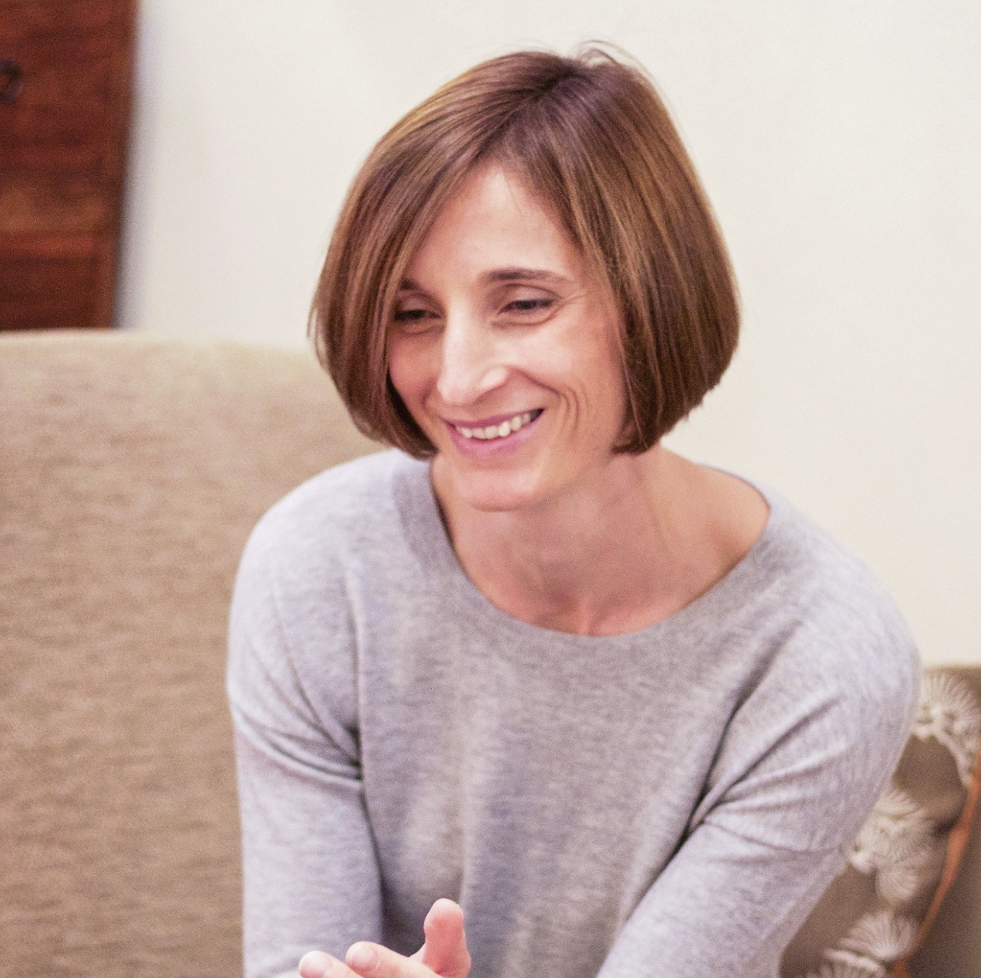A woman with short, light brown hair smiling, wearing a light grey sweater, sitting indoors on a beige sofa with a wooden surface in the background.