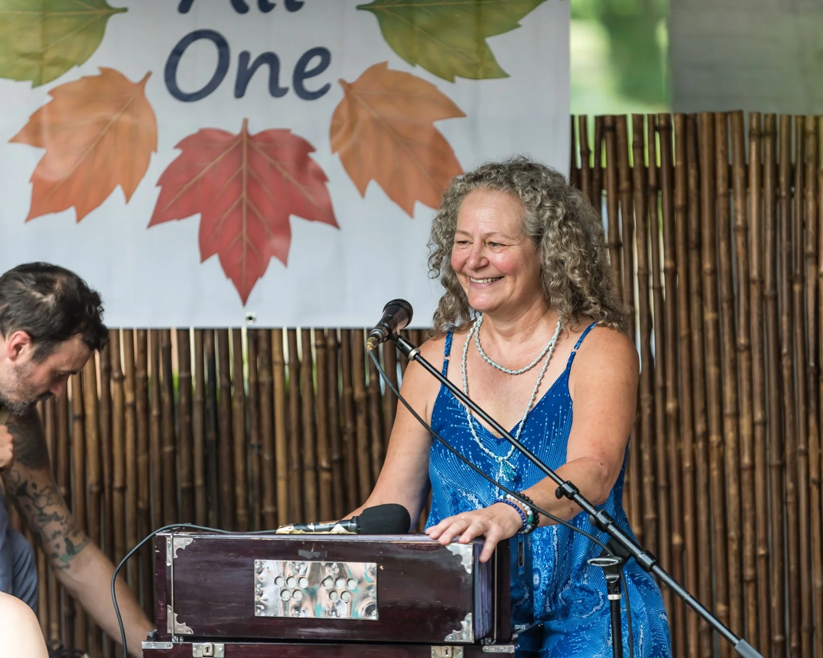 A woman in a blue dress smiling while standing at a keyboard, with a microphone in front of her and a person with tattoos on the left side, against a backdrop of a banner with autumn leaves and the word "One".