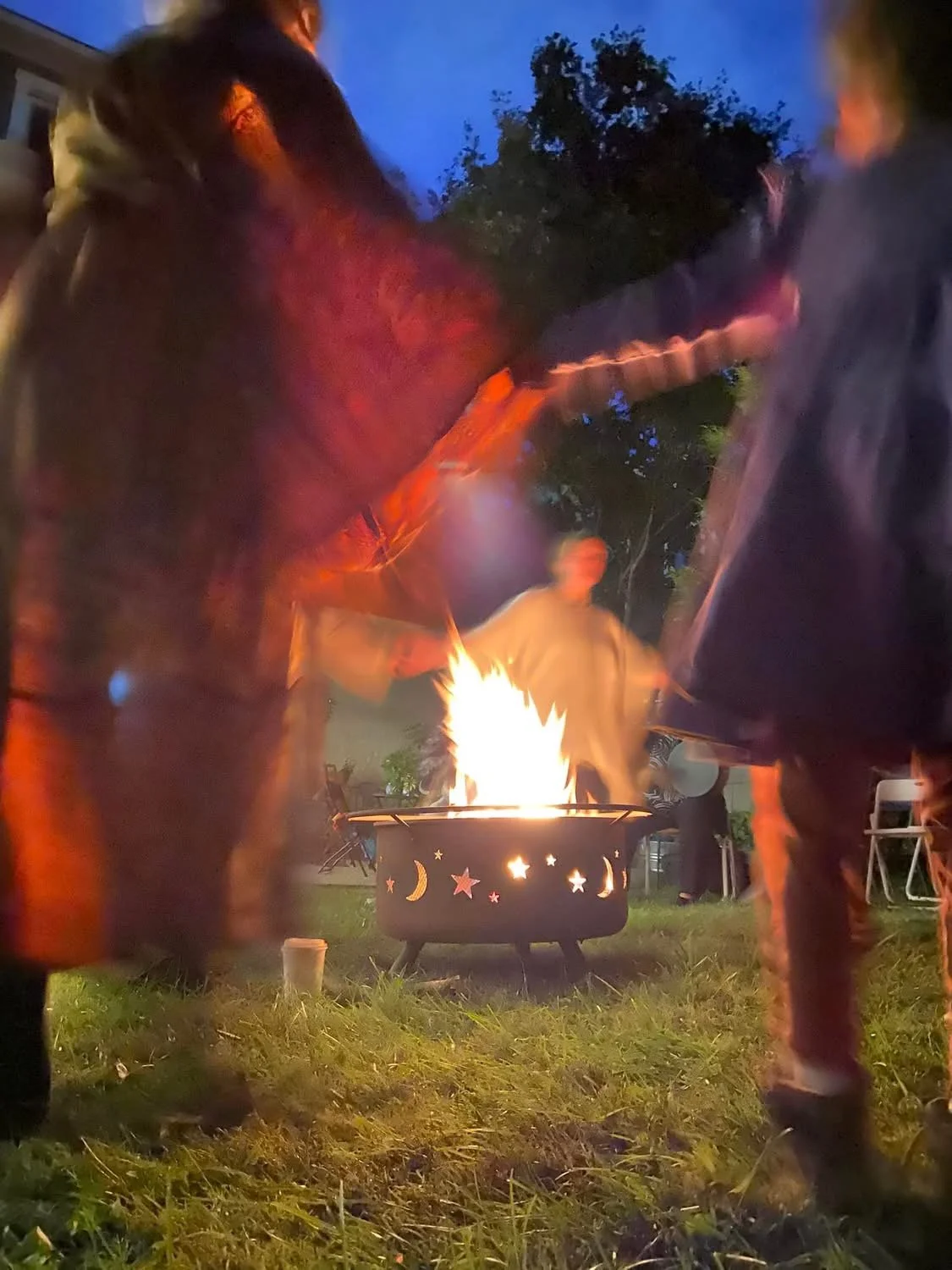 People gathered around a fire pit with moon and star cutouts, outdoors at dusk, some dancing or moving to music, trees in the background.