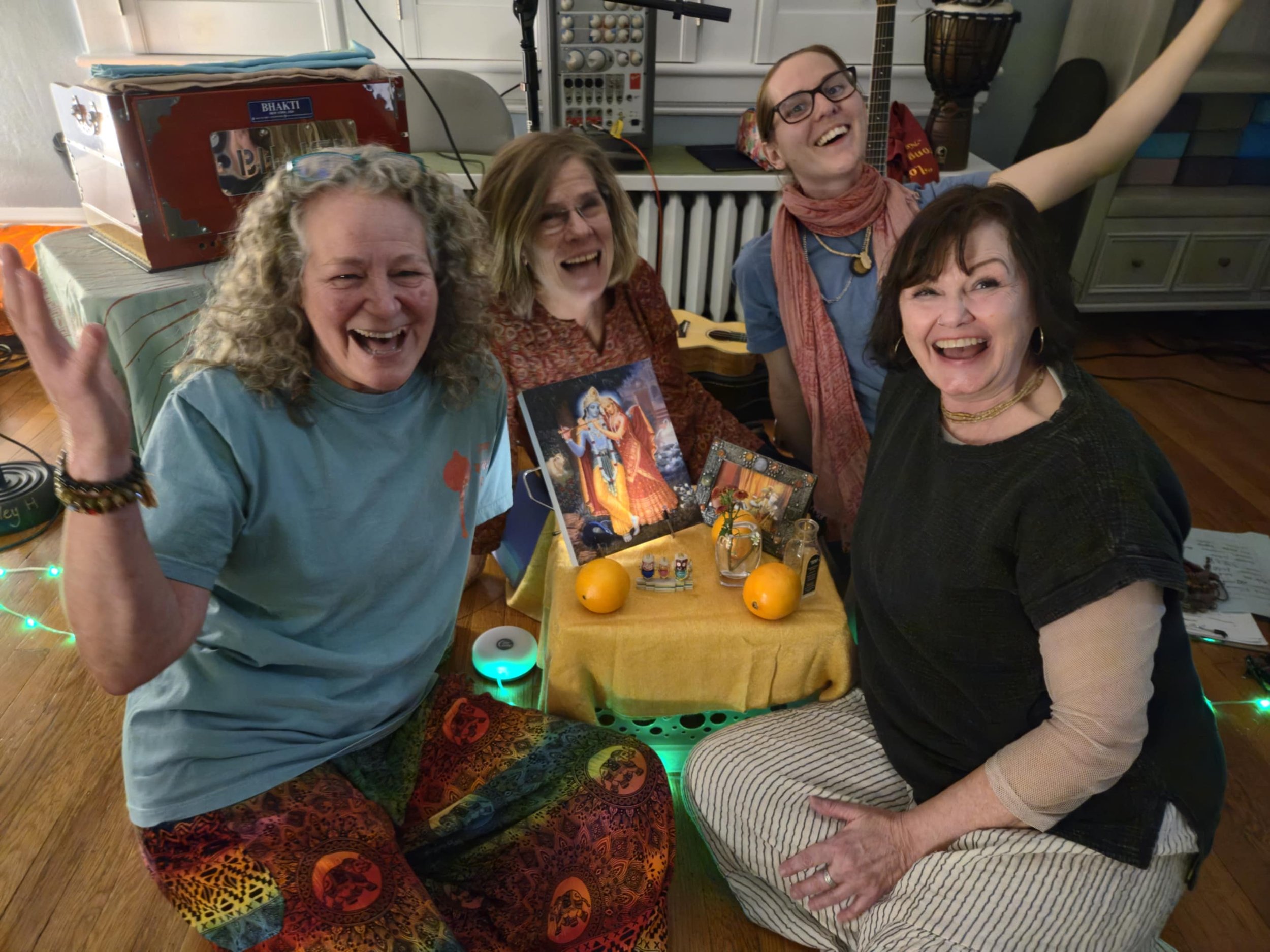 Four women smiling and laughing together around a decorated table with books and oranges, in a room with musical instruments and electronics in the background, celebrating or having a gathering.