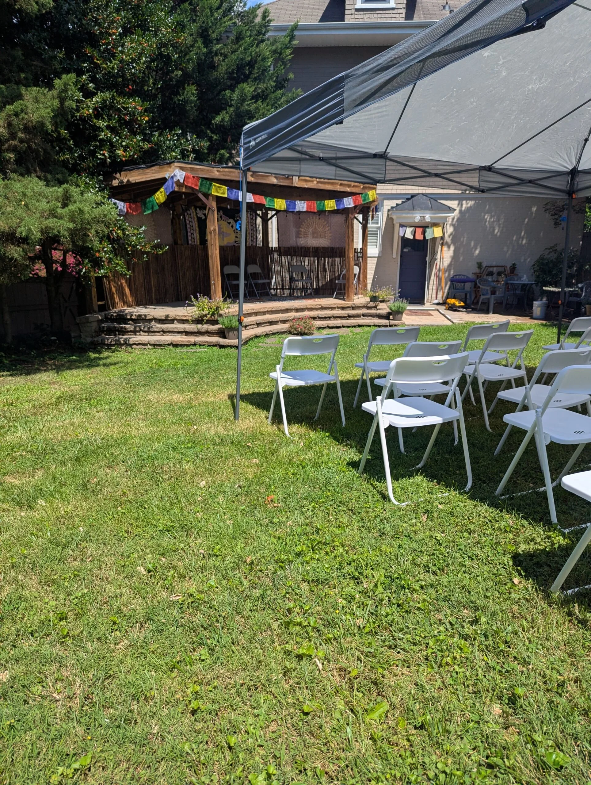A backyard setup for an outdoor event with white folding chairs arranged in rows on a grassy lawn, a large canopy tent, and a small stage area decorated with colorful flags and prayer flags, surrounded by trees and plants.