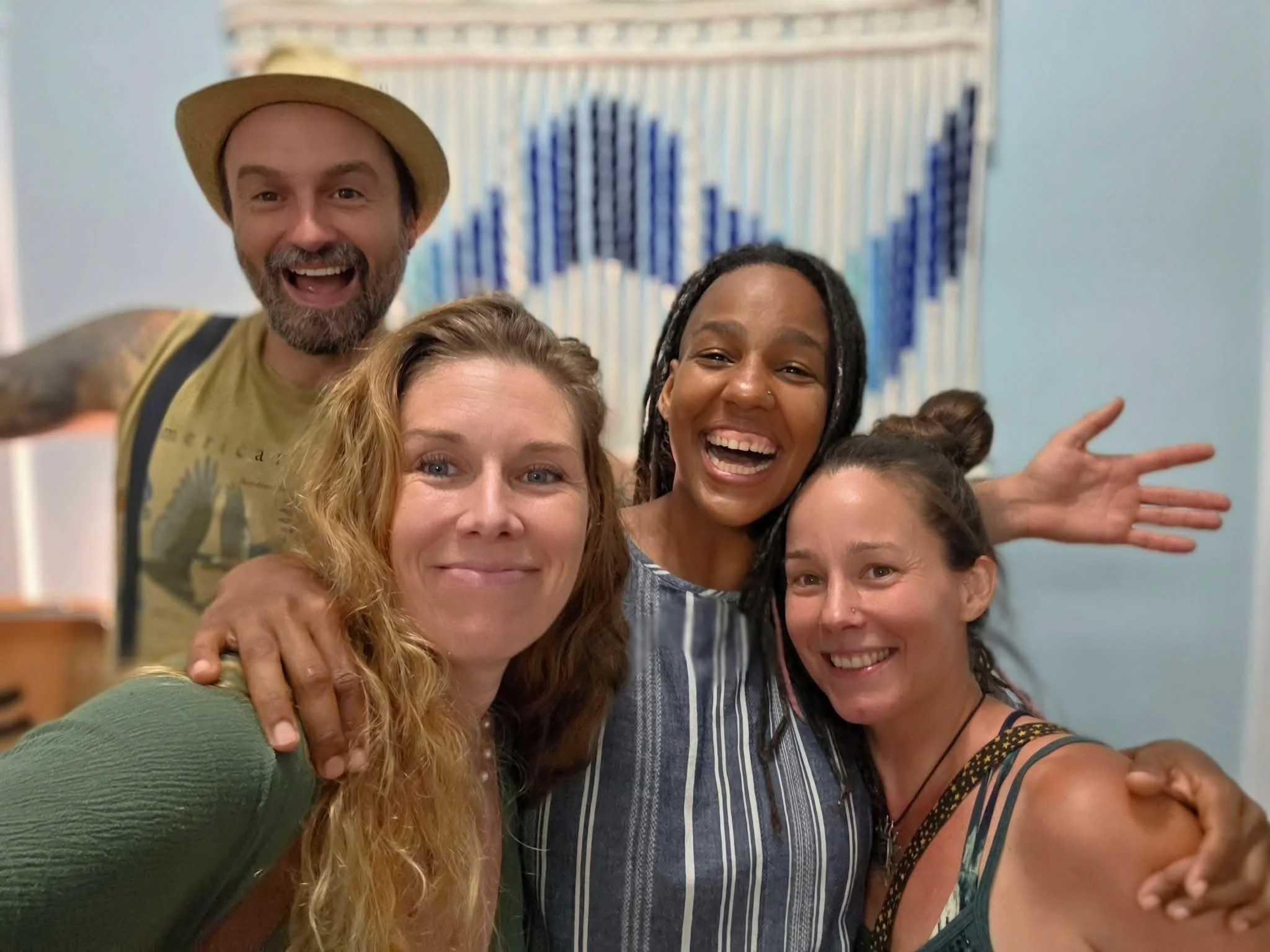 A group of six smiling people taking a selfie together indoors, with a colorful artwork on the wall behind them.