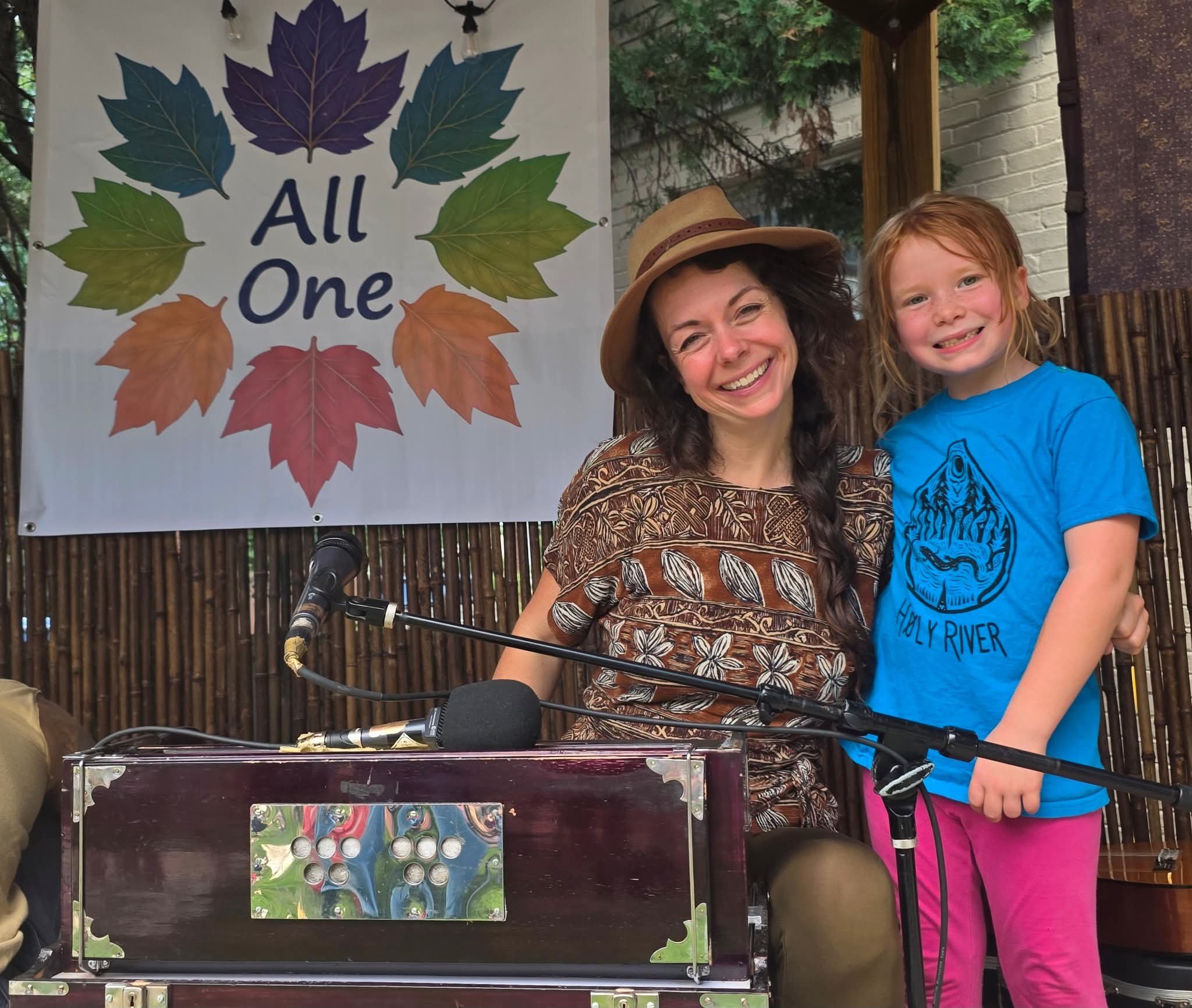 Smiling woman wearing a hat and a young girl with red hair, both standing next to each other in front of a sign with colorful autumn leaves and the words 'All One.' They are outdoors, and there is a microphone stand in front of them.