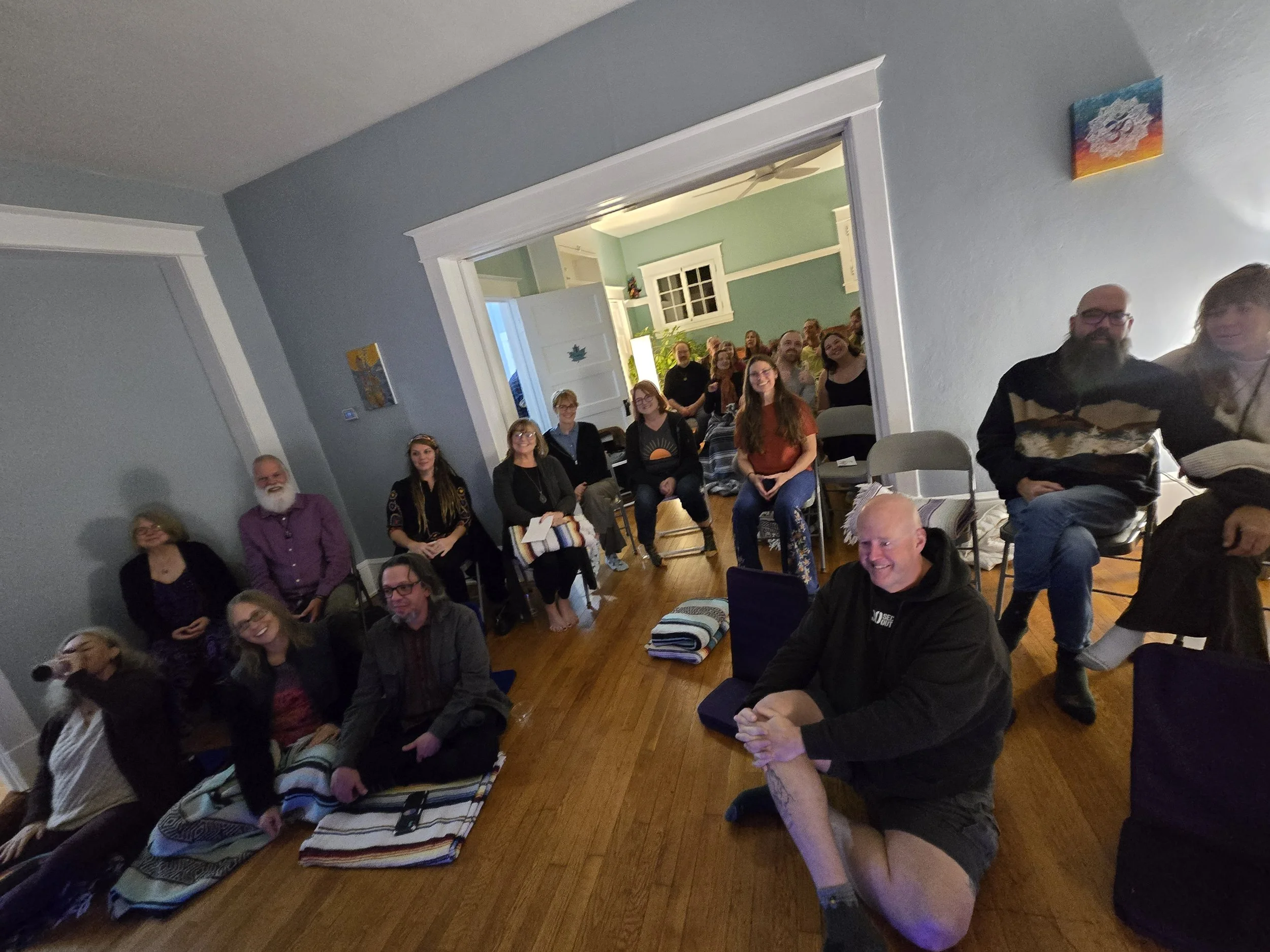 A group of people gathered indoors, seated and standing, smiling and looking towards the camera in a cozy, well-lit room with artwork on the walls and a doorway leading to another room.