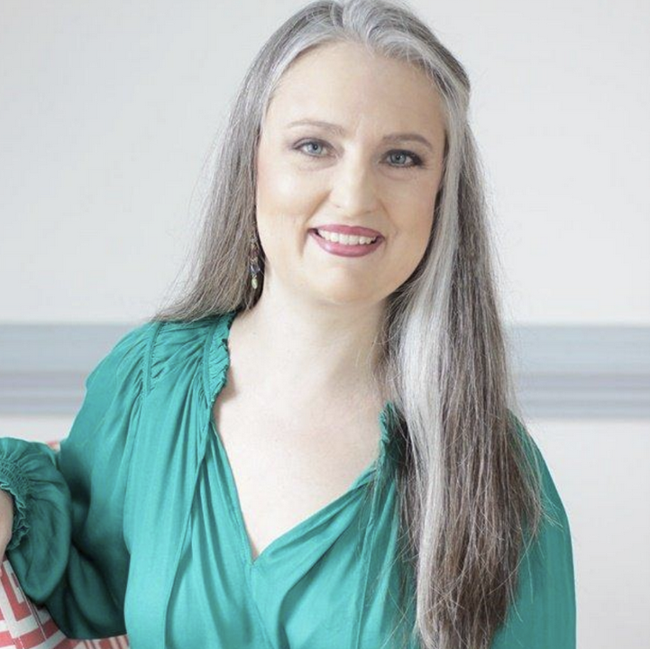 A woman with long gray hair, wearing a teal blouse, smiling at the camera.