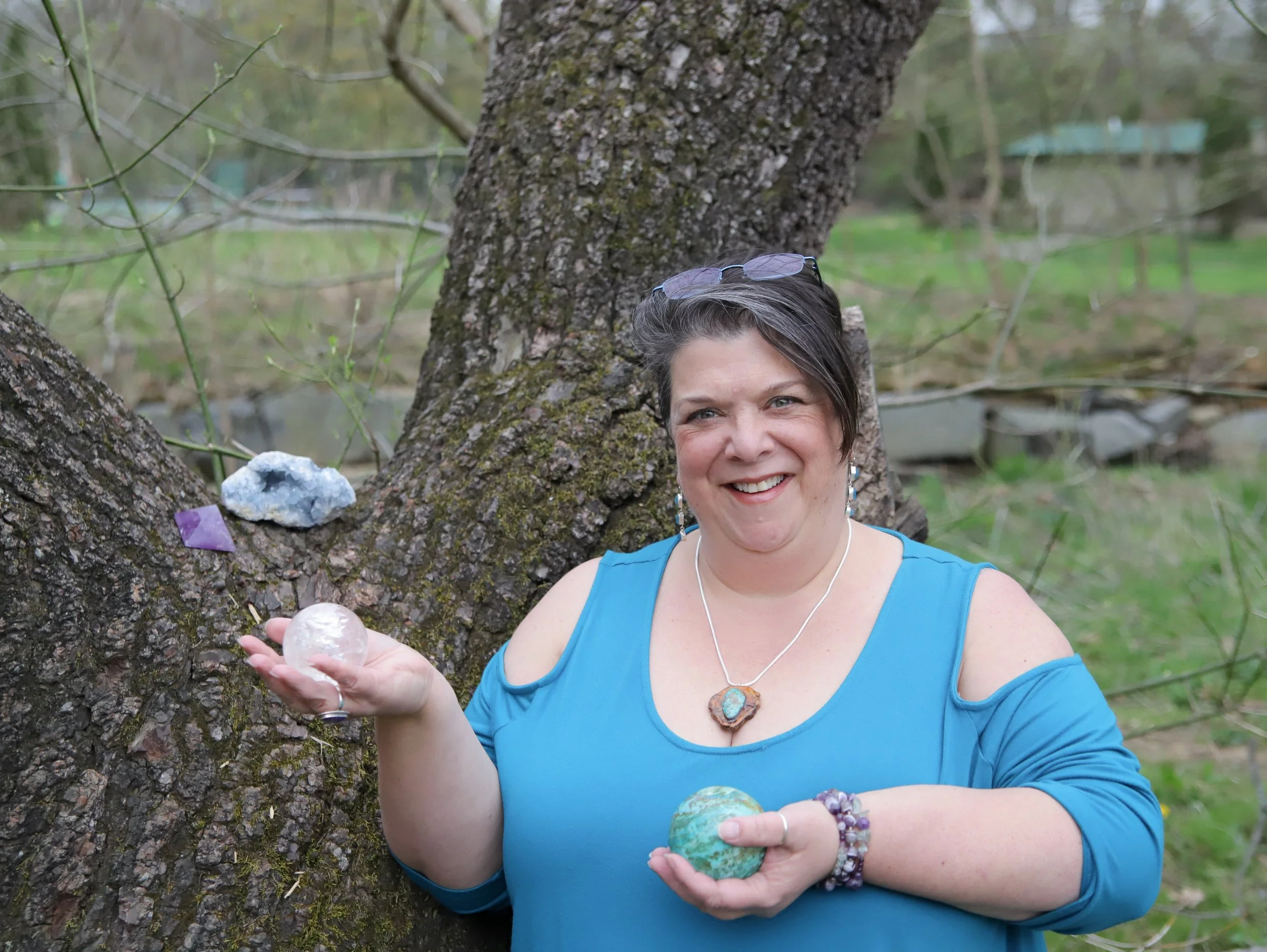 A woman in a blue top standing outdoors in front of a large tree, holding a large crystal ball and wearing jewelry including a necklace and bracelets, with glasses on her head and crystals on the tree.