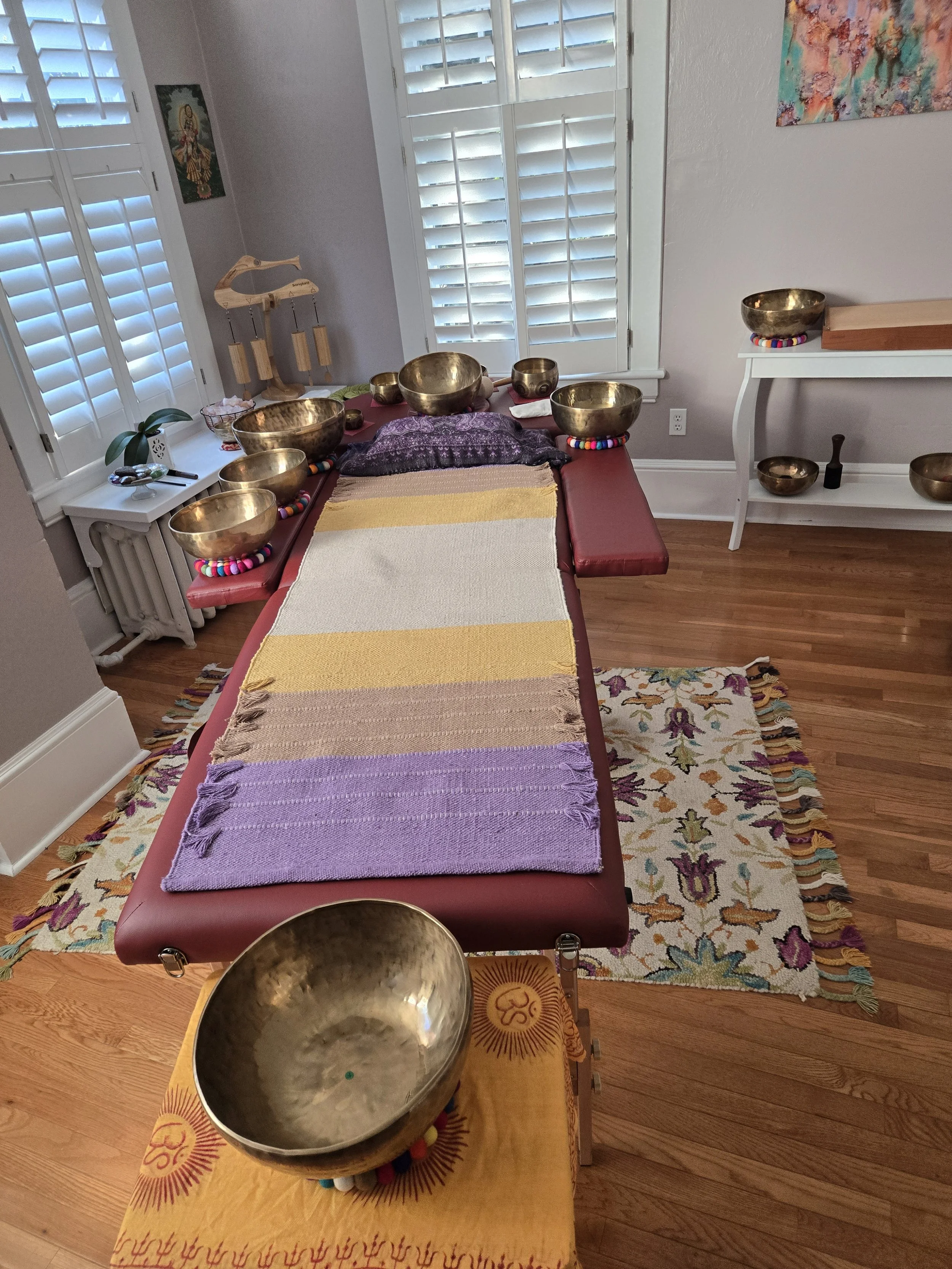 This image shows a healing or meditation space with a massage table covered with colorful blankets and pillows, several brass singing bowls on the table and a floor, a small altar with a yellow cloth featuring a Sun symbol, and a window with white sh