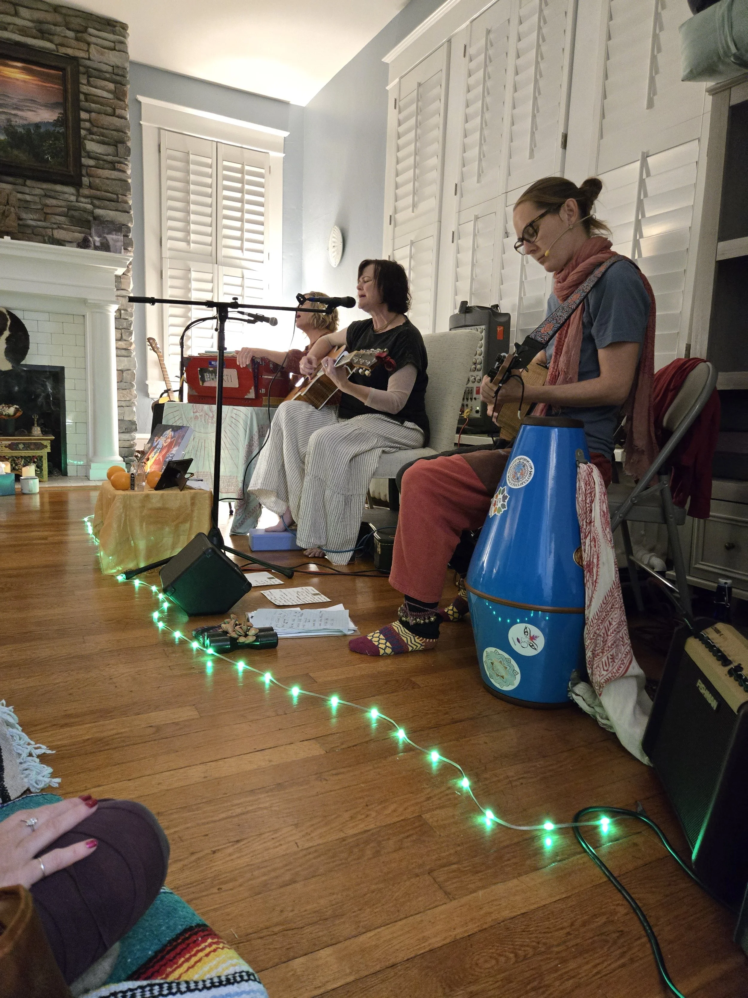 Three women are sitting on chairs with musical instruments, singing and playing guitar and other instruments in a room with white walls and shutters, decorated with string lights and other items.