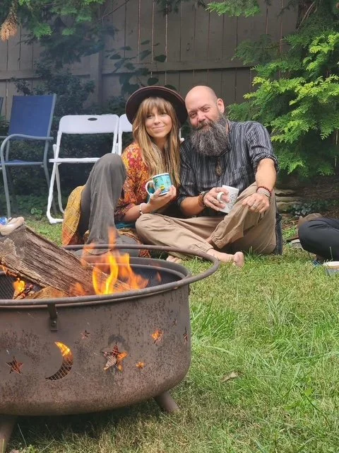 Two people, a woman and a man, sitting on grass near a campfire in a backyard, holding mugs and smiling. There are chairs and greenery in the background.
