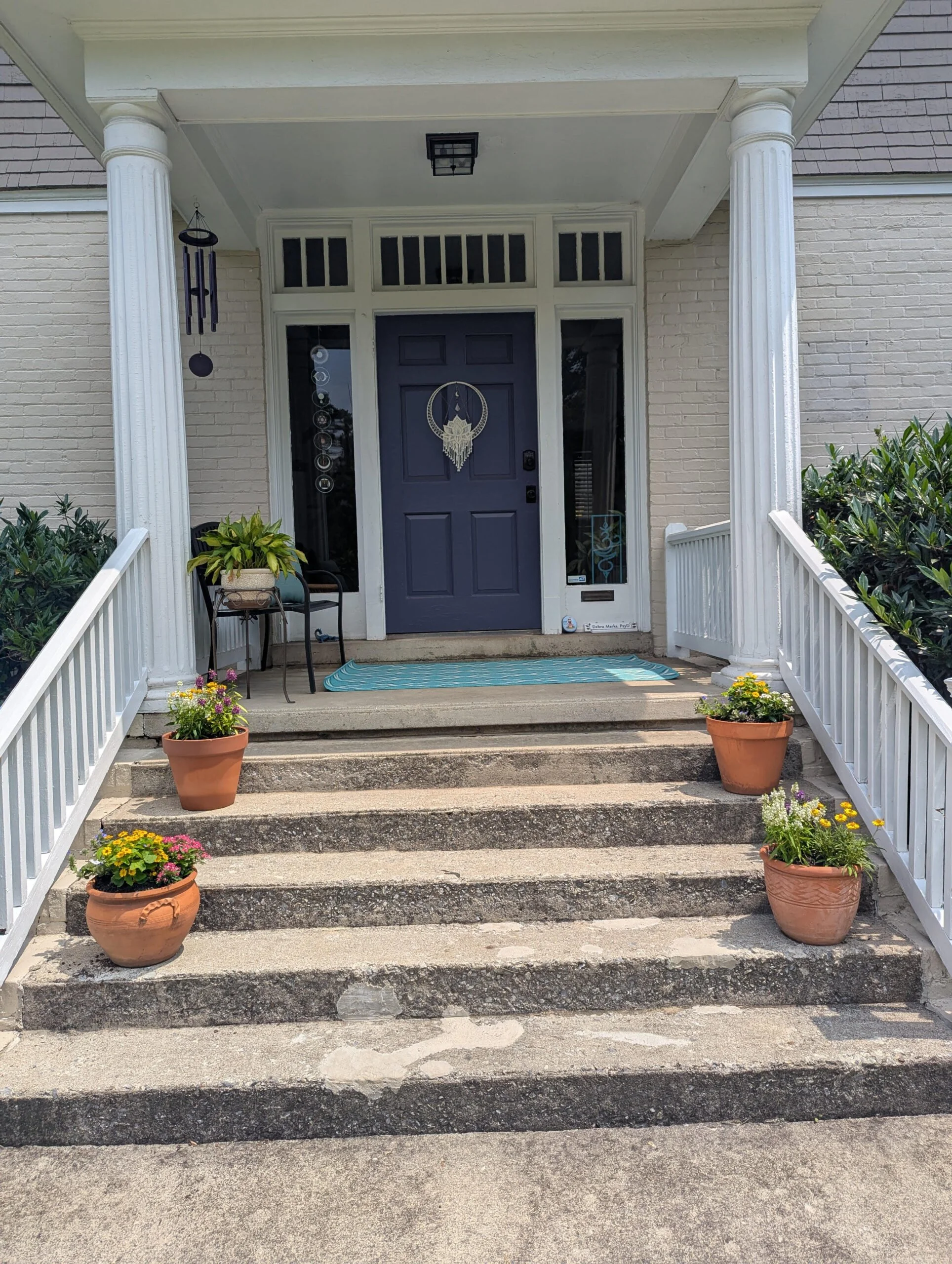 Front porch with concrete steps, white railing, potted flowers, plants, and a dark blue door decorated with a circular dreamcatcher. Two white columns support the porch roof, and a wind chime hangs on the left side of the porch.