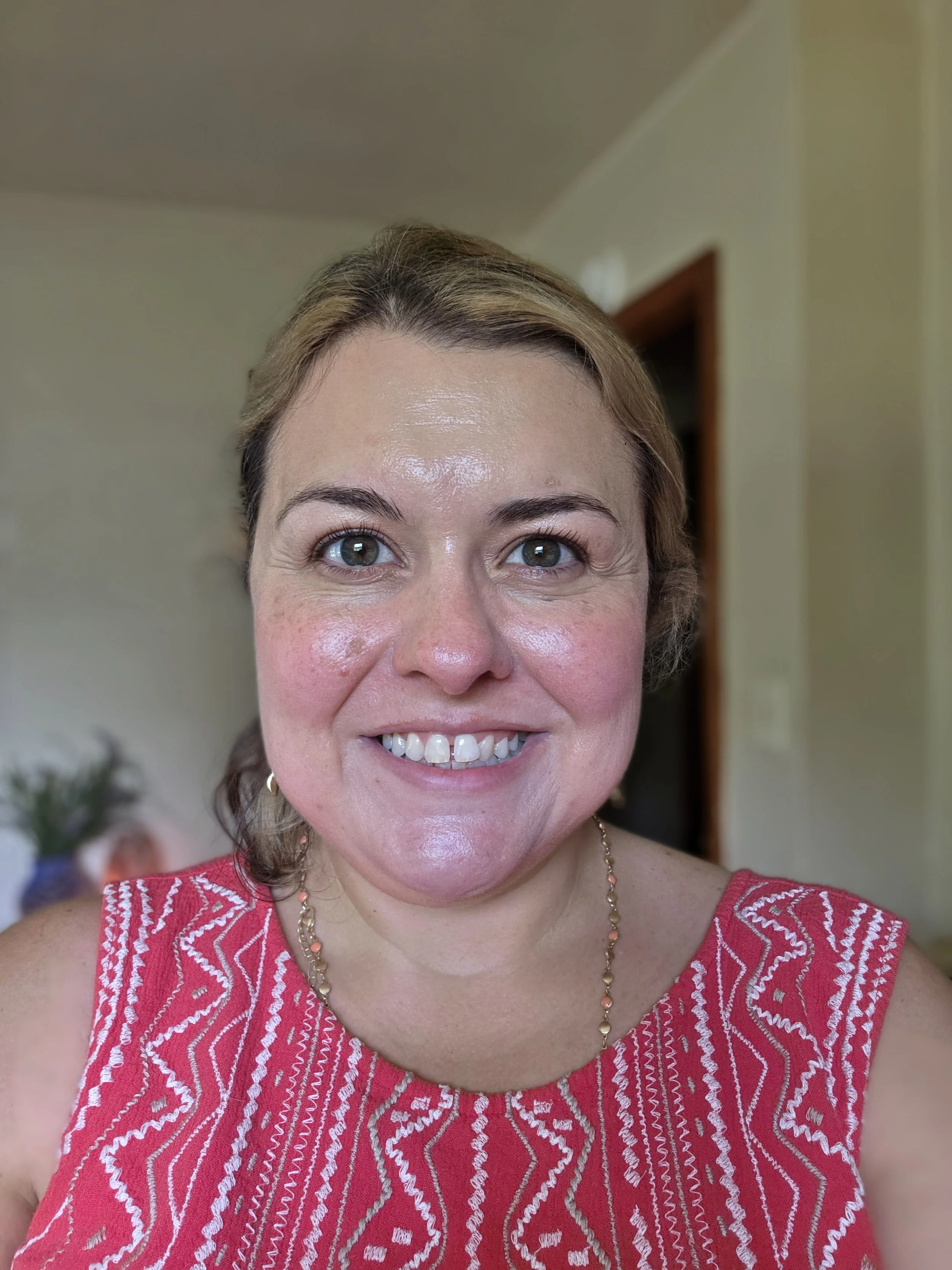 Close-up of a woman with short brown hair, wearing a red patterned top, smiling, in an indoor setting with beige walls and a doorway in the background.