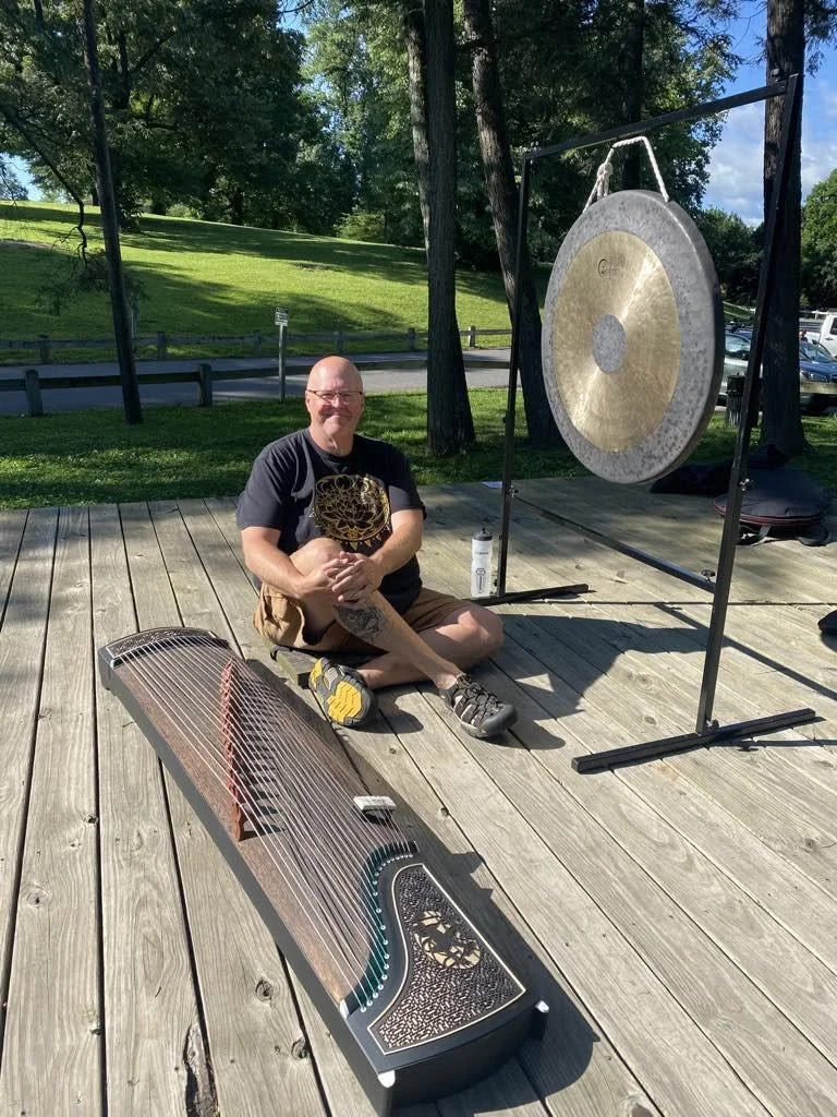A man sitting cross-legged on a wooden deck outdoors next to a large gong hanging from a metal frame and a harp-shaped stringed instrument on the deck.