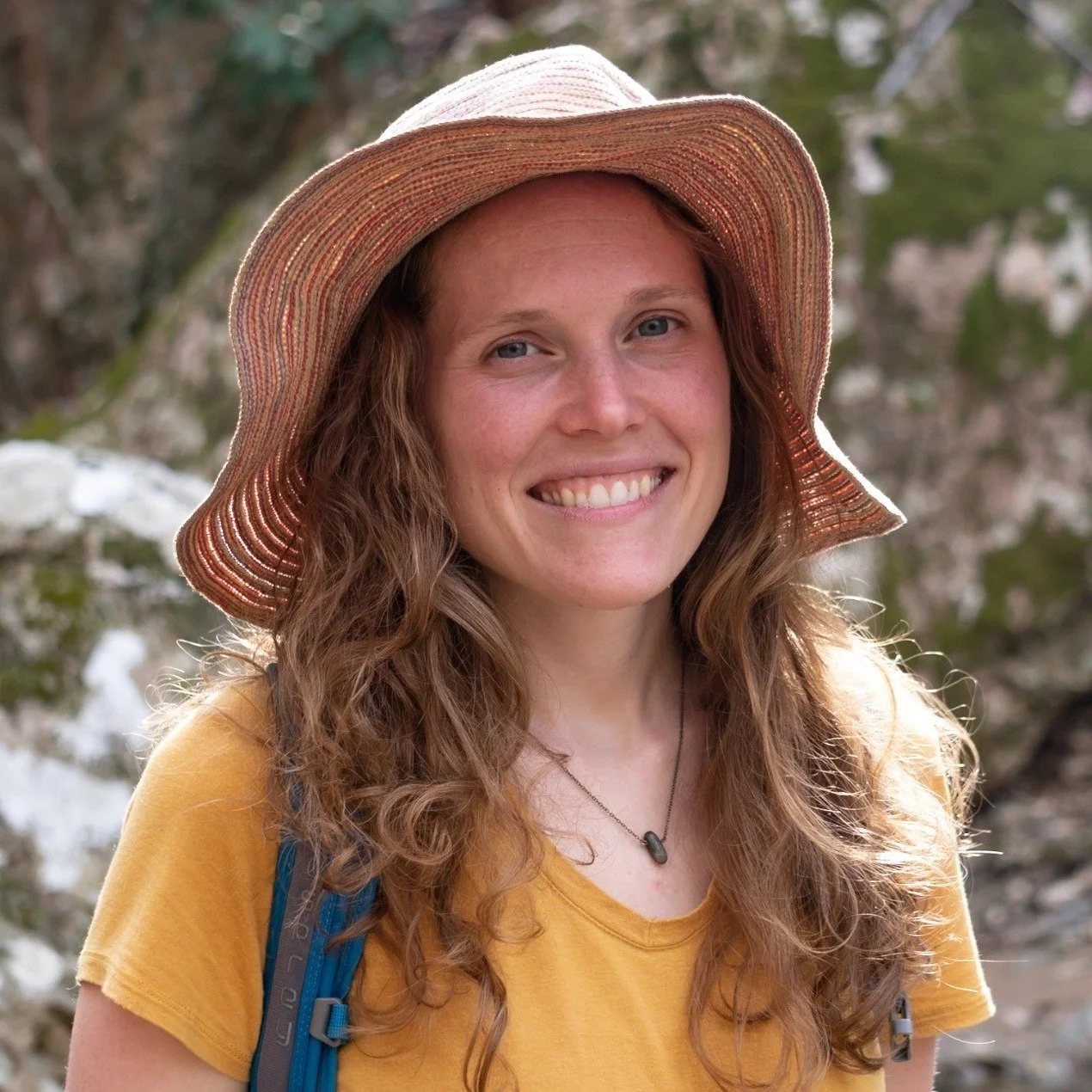 A smiling woman with long curly hair wearing a wide-brimmed straw hat and yellow t-shirt, outdoors with rocky and green background.
