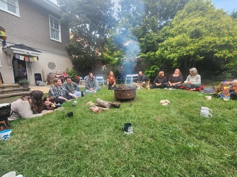 Group of people sitting on the grass around a fire pit in a backyard, with trees and a house in the background.