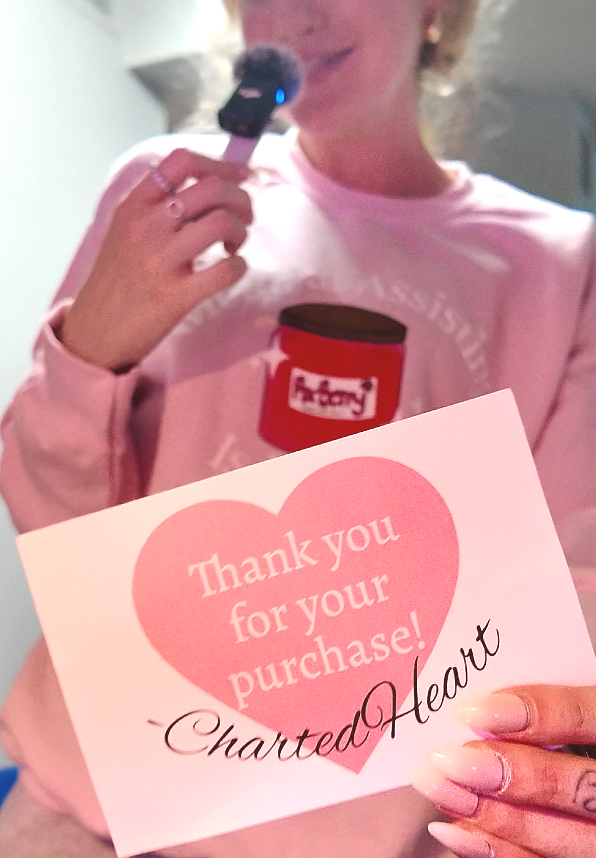 A woman in a pink shirt holding a small jar of jelly and a pink card with a heart that says, 'Thank you for your purchase!'. She is smiling and her face is partially visible.