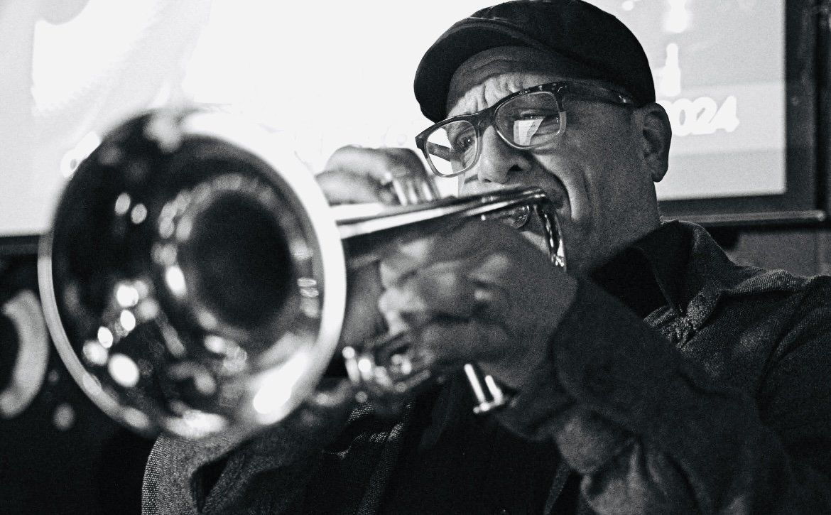 A man wearing glasses and a cap playing a trumpet in a dimly lit setting.