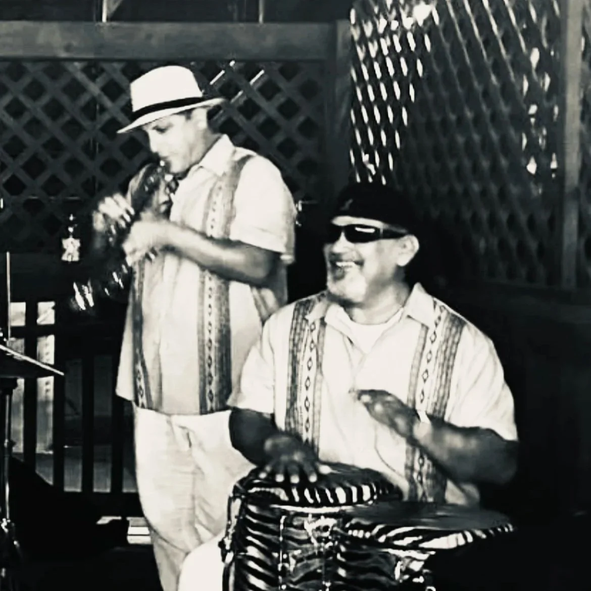 Two men wearing sunglasses and Cuban-style shirts with patterns, one playing conga drums, one playing trumpet, both smiling, in an outdoor setting with lattice wood fencing.