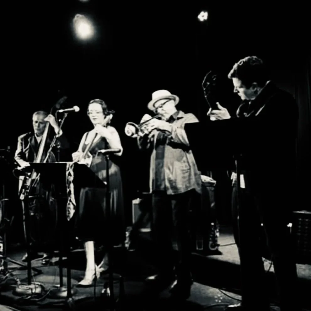 Black and white photo of five musicians playing on stage, including a saxophonist, a female singer, a clarinet player, and two other musicians with their instruments, under stage lighting.