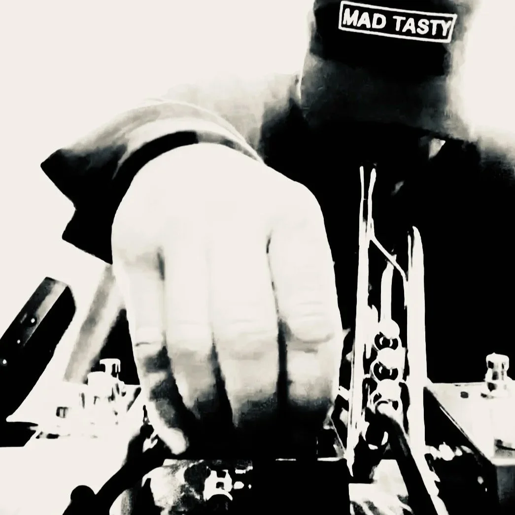 A person wearing a hooded sweatshirt with a 'MAD TASTY' patch on the hood, working with a guitar or bass guitar on a workbench or desk, in a monochrome, high-contrast style.