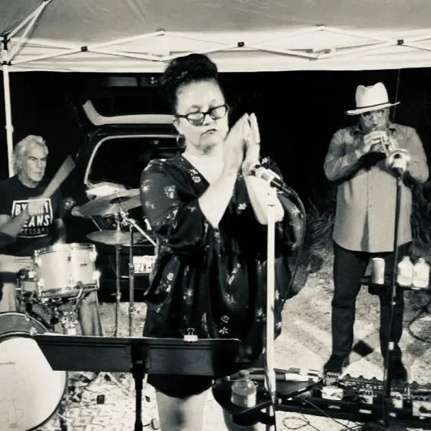 Black and white photo of three musicians performing at night under a canopy. A woman in the center is singing or praying with her eyes closed and hands together. To her left, a man is playing drums, and on her right, a man in a hat is playing a trumpet.