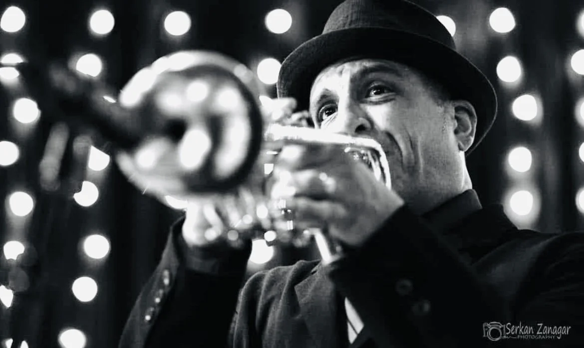A man playing the trumpet, wearing a hat, with a blurred background of bright lights.