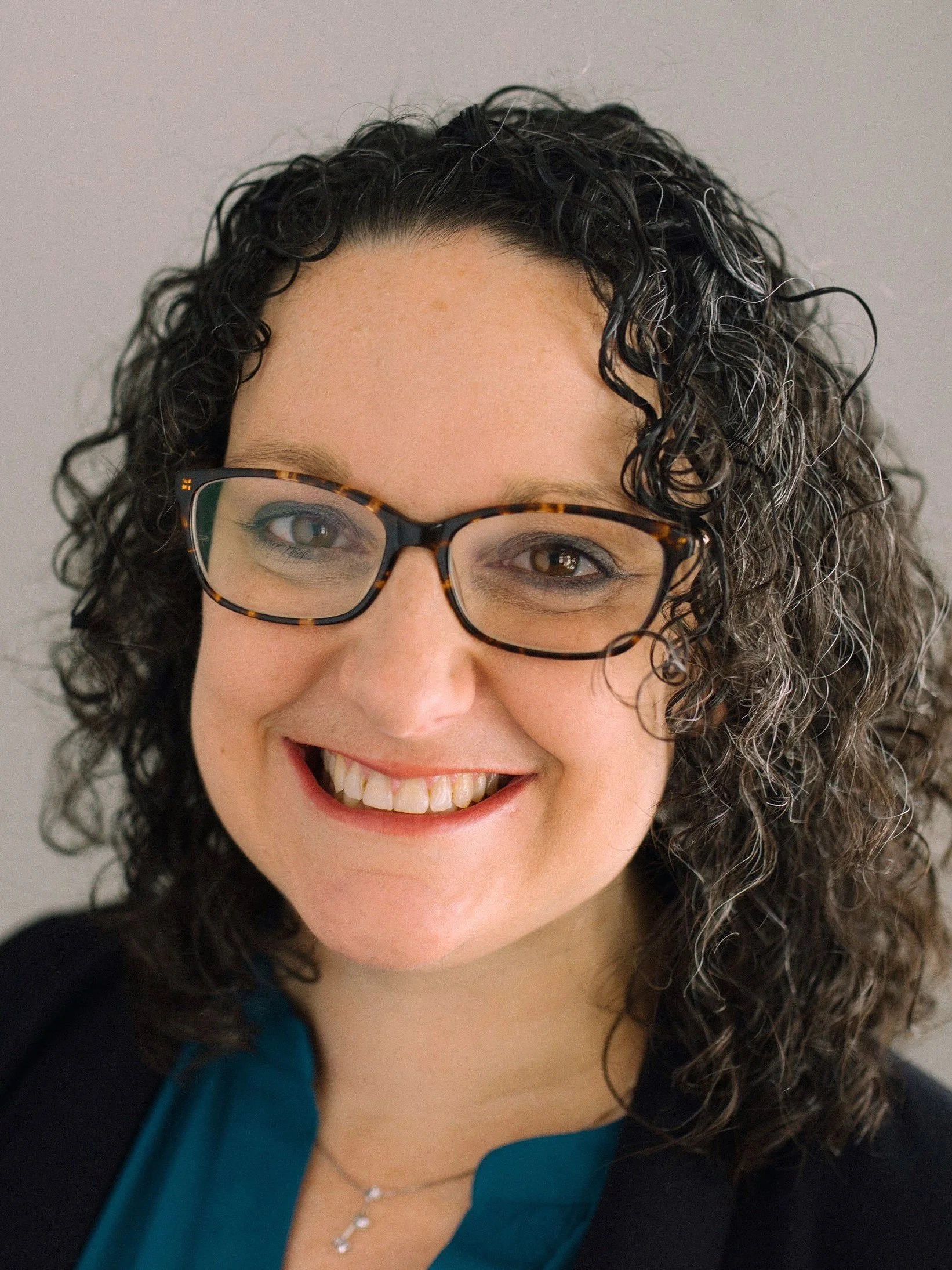 A woman with curly black hair smiling, wearing glasses with tortoise-shell frames and a blue top.