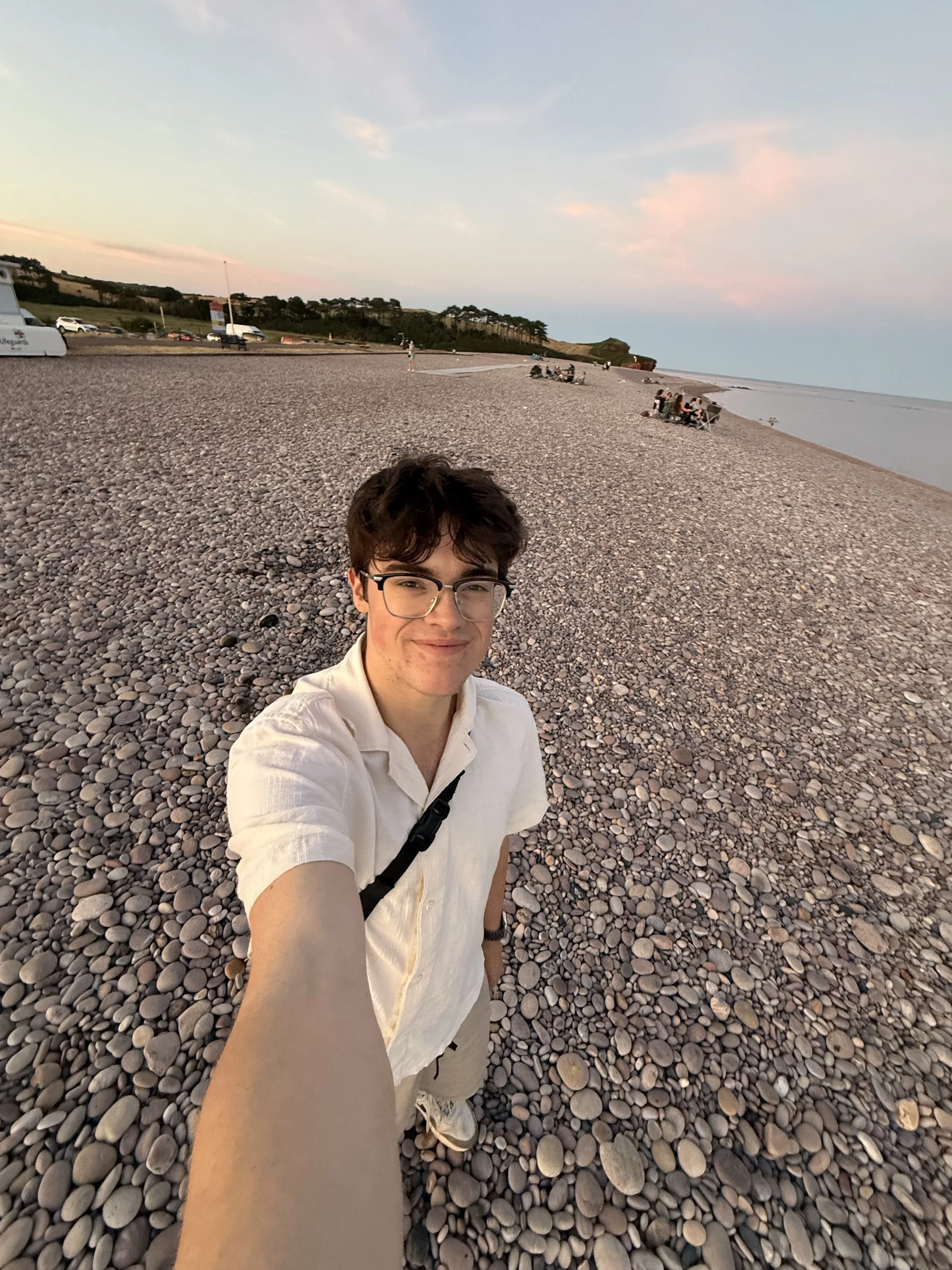 Young man with glasses taking a selfie at a pebble-covered beach during sunset, with a few people sitting and walking along the shore, and a grassy hillside in the background.