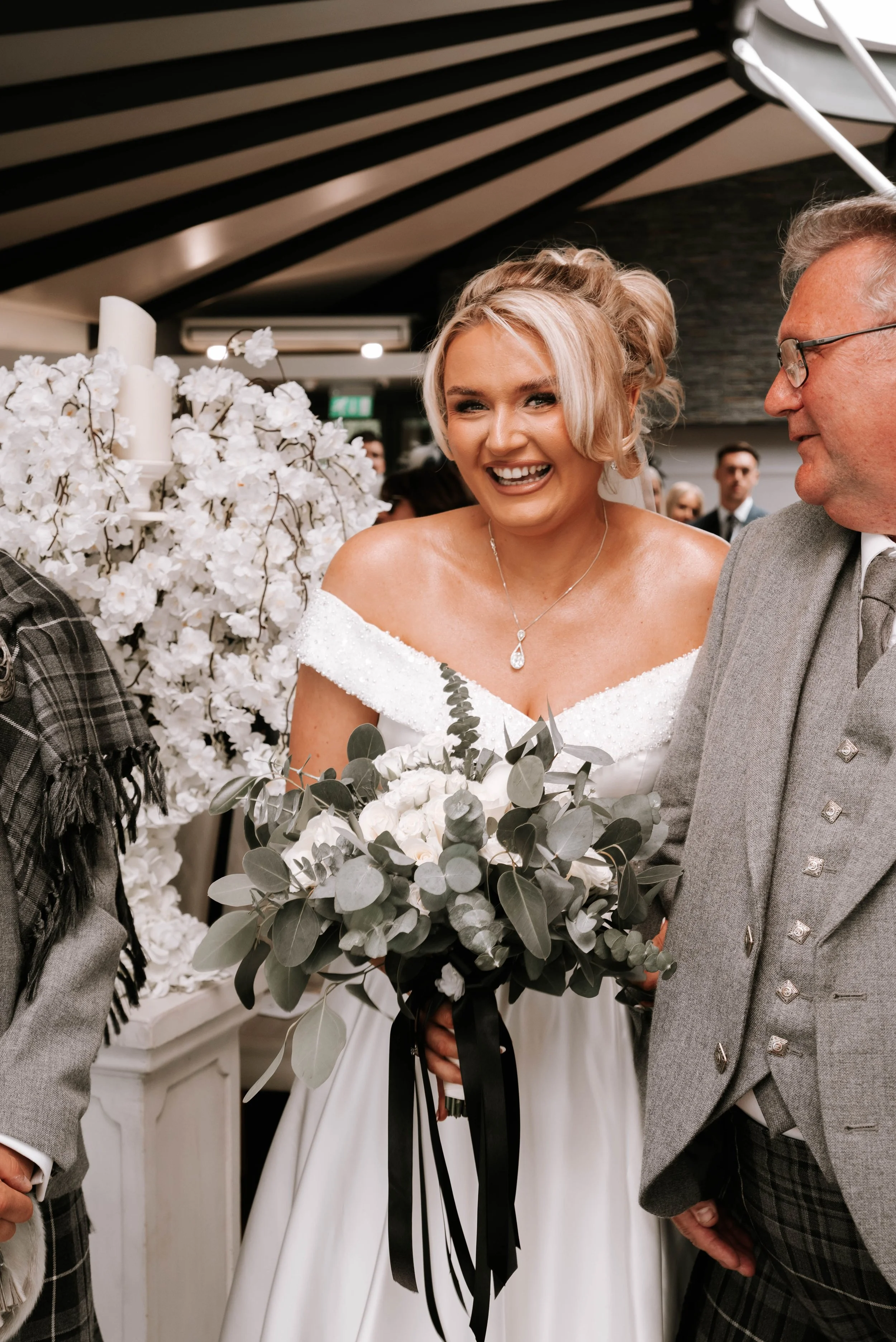 A woman wearing a wedding dress smiling with a man in a gray suit holding a bouquet of white flowers and eucalyptus leaves at a wedding ceremony.