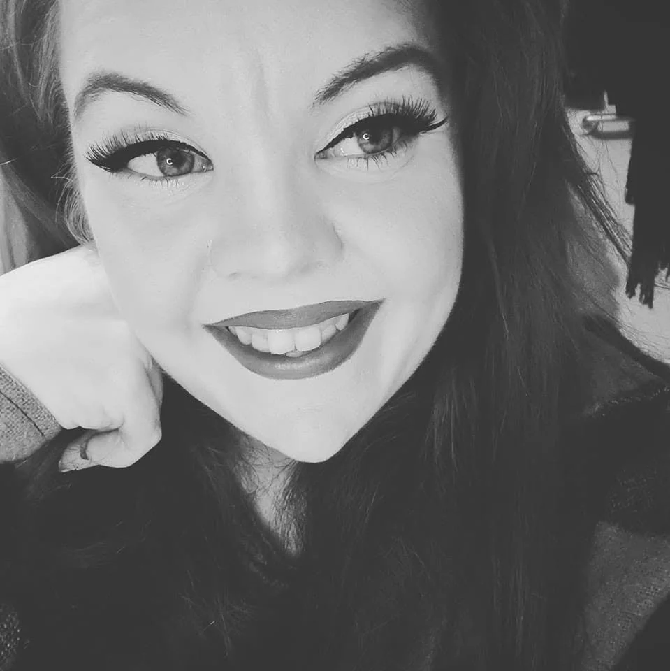 Black and white close-up photo of a woman smiling with long dark hair, wearing makeup with prominent eyelashes and lipstick, resting her chin on her hand.