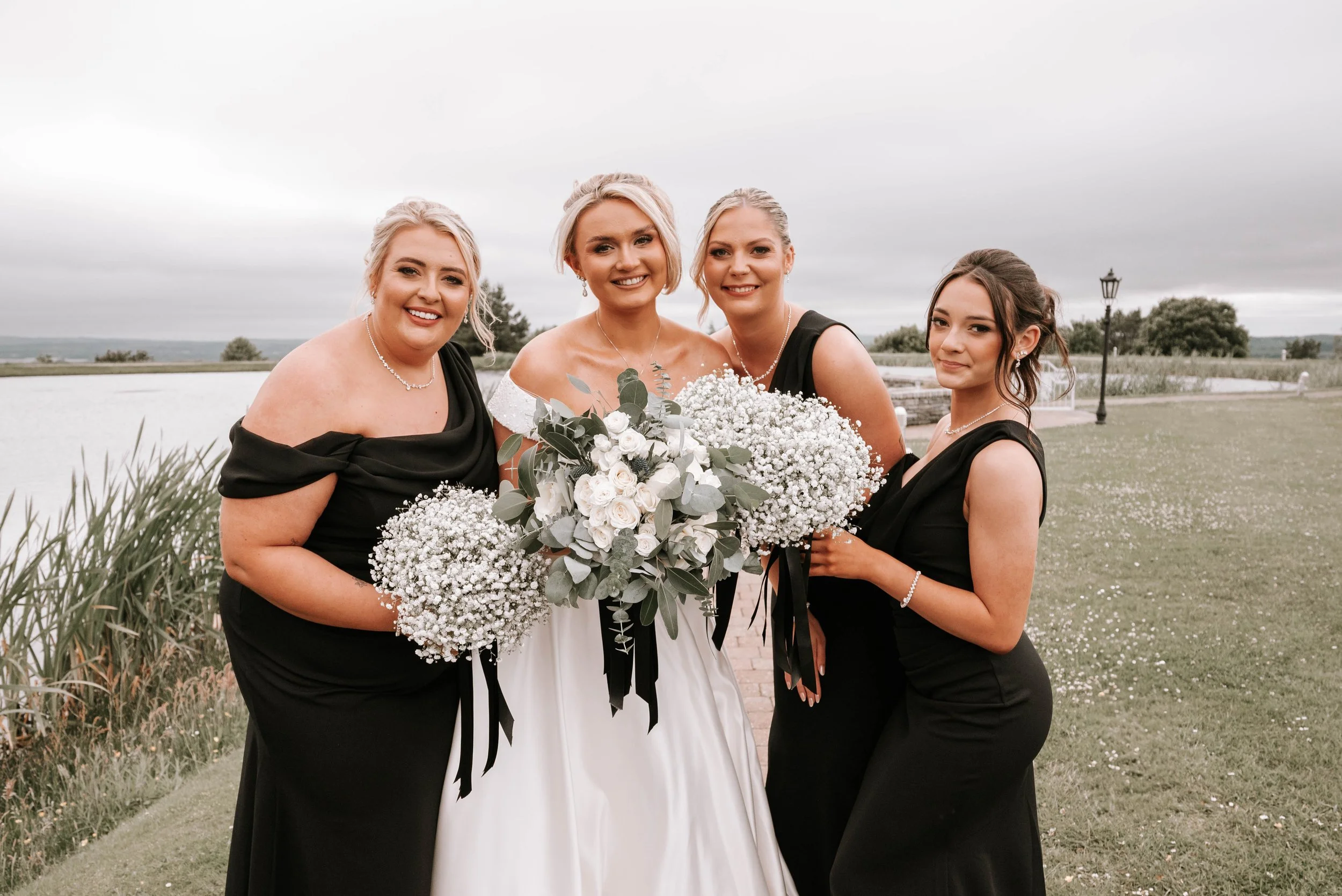 Four women, dressed in formal attire, standing outdoors by a lakeside on a cloudy day. The central woman is wearing a white dress and holding a large bouquet of white and green flowers, indicating a wedding or special event. The other women are weari