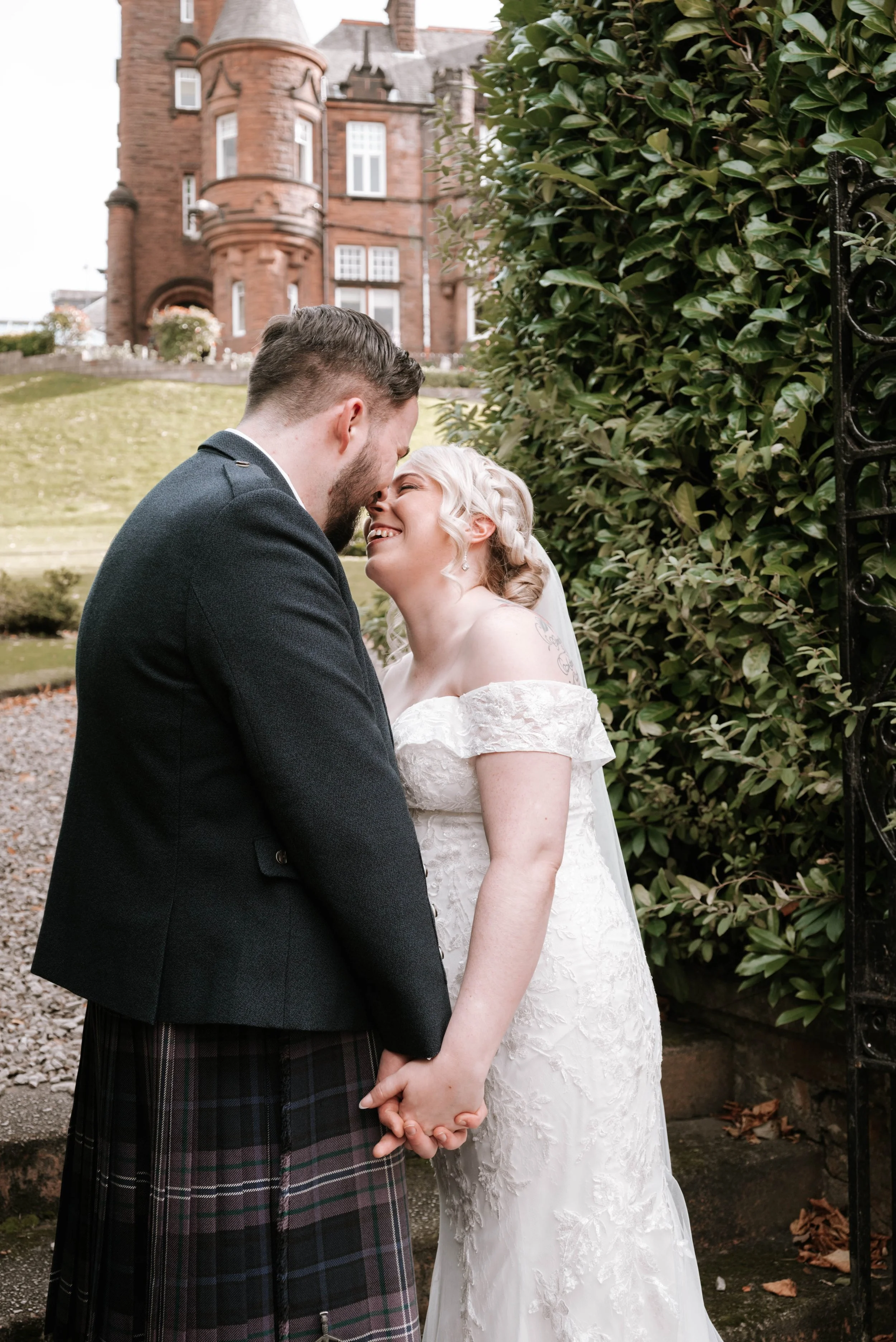 A bride and groom holding hands and touching foreheads in a garden, with a large brick castle in the background.