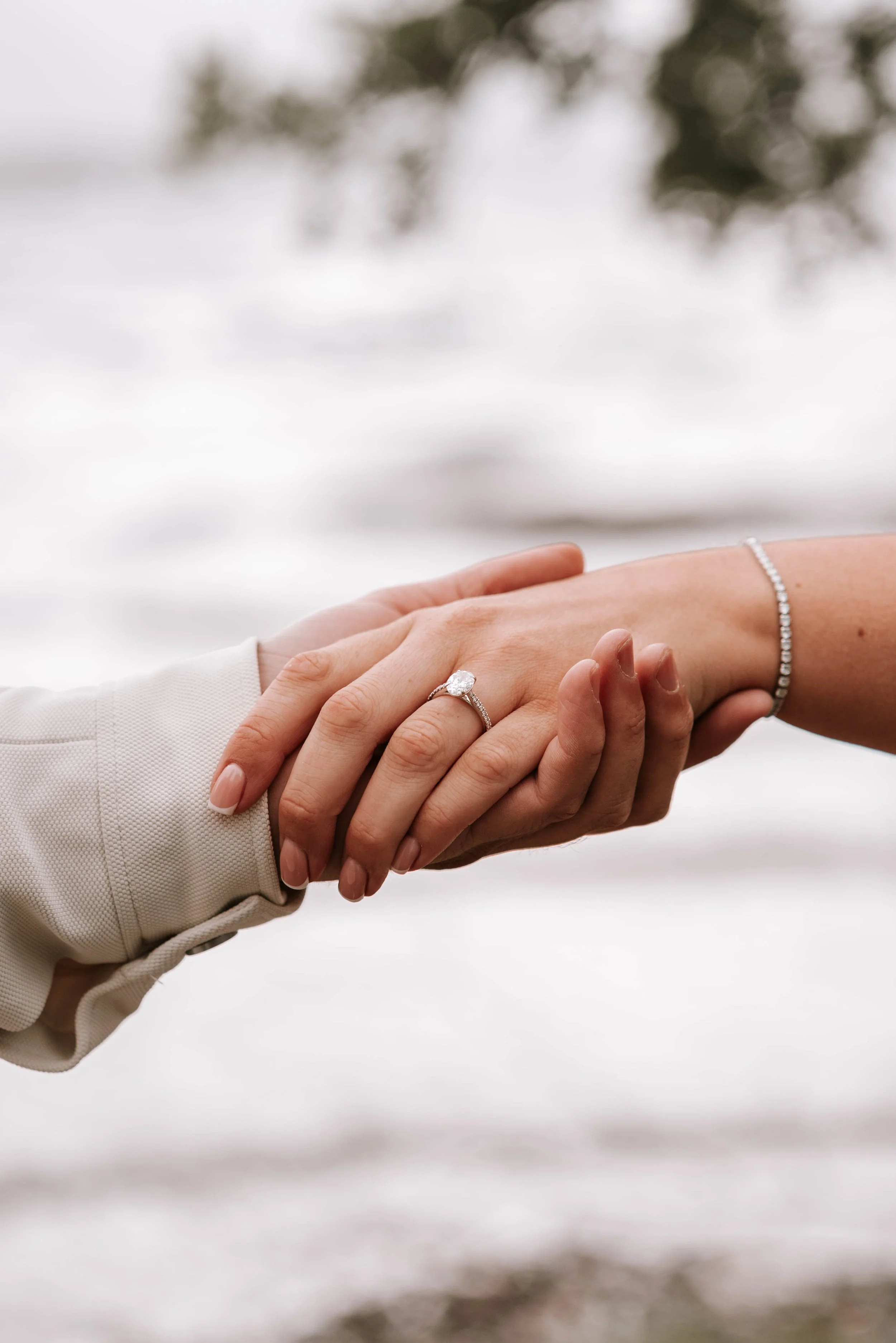 Close-up of two hands holding each other; one hand has an engagement ring, and the other has a bracelet. The background shows a snowy landscape with blurred trees.