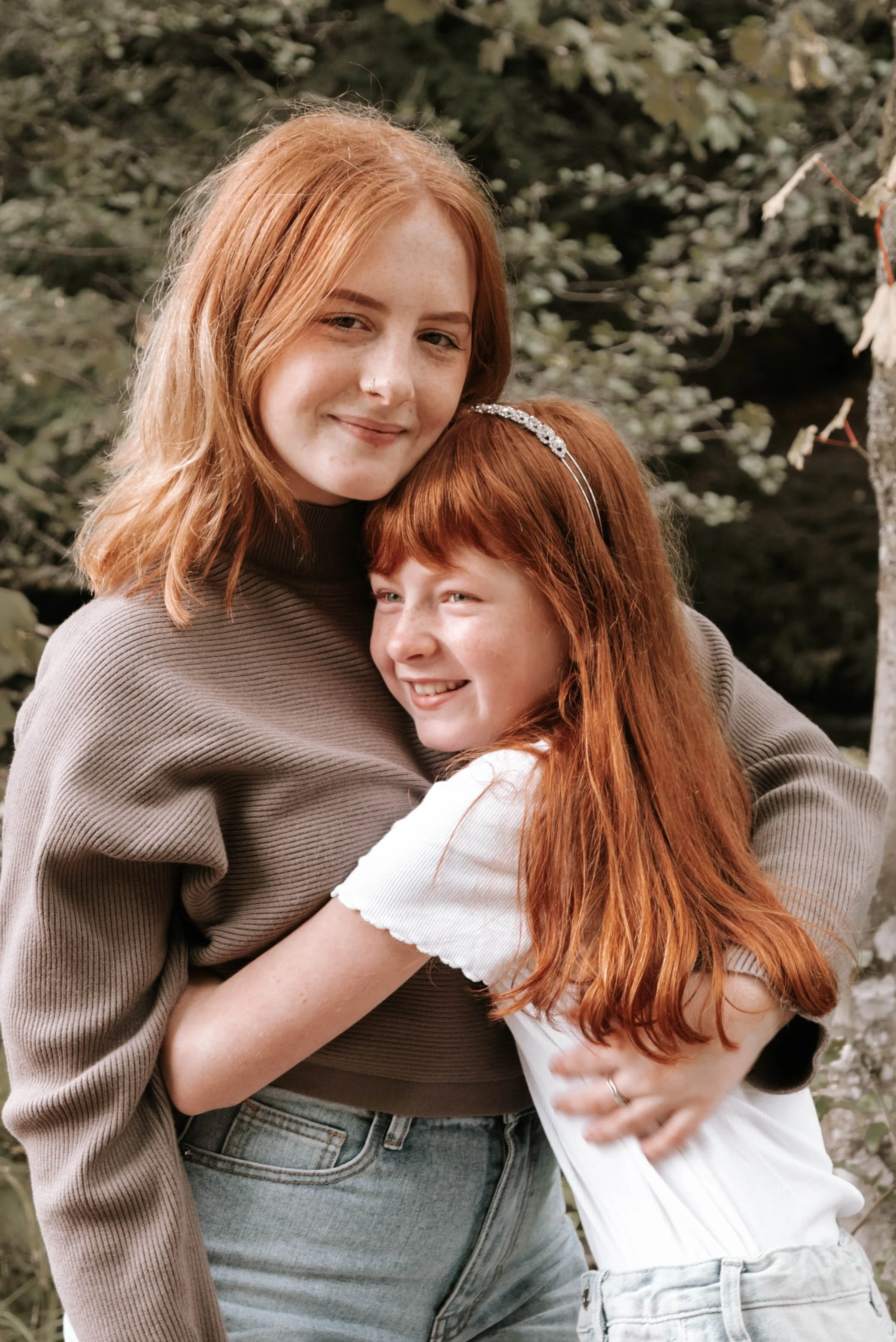 Two young women with red hair embracing outdoors, smiling, against a background of trees and foliage.