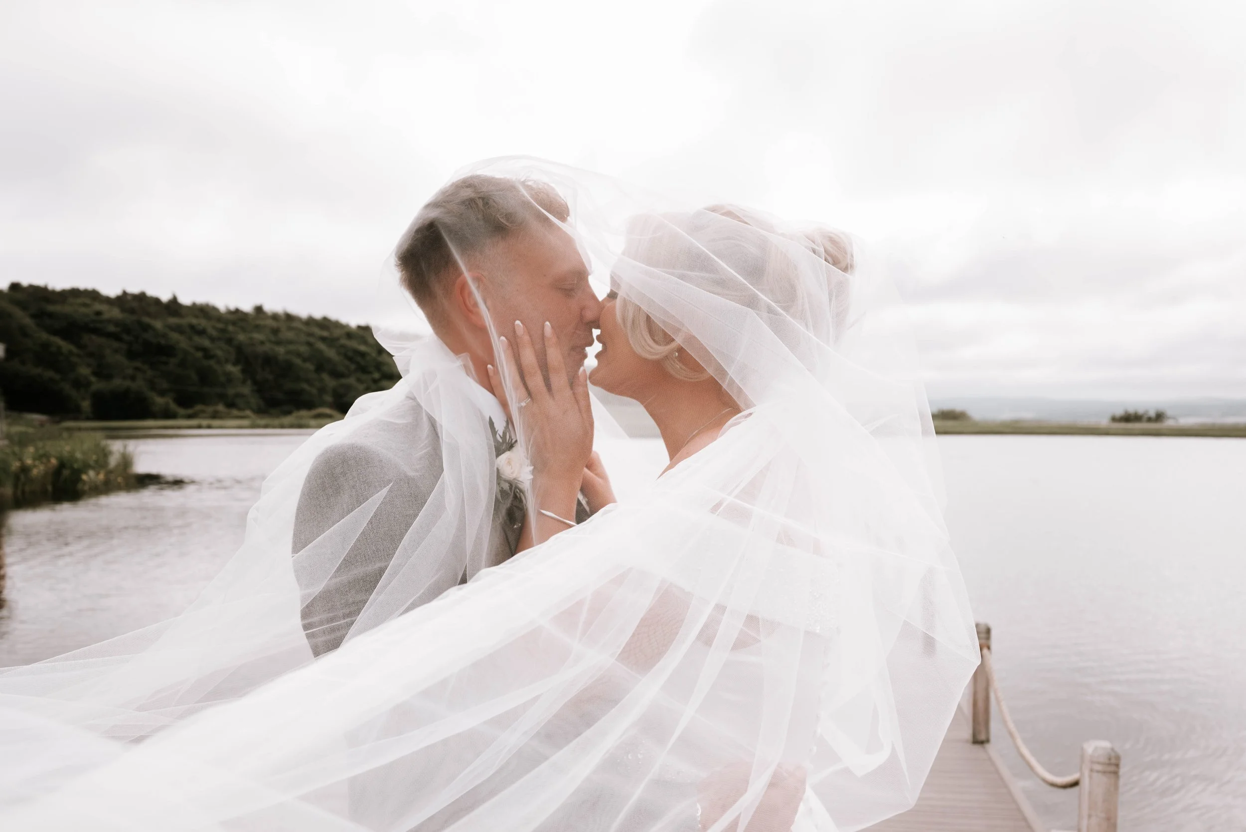 A couple dressed in wedding attire sharing a kiss under a veil near a body of water with a forested hillside in the background.