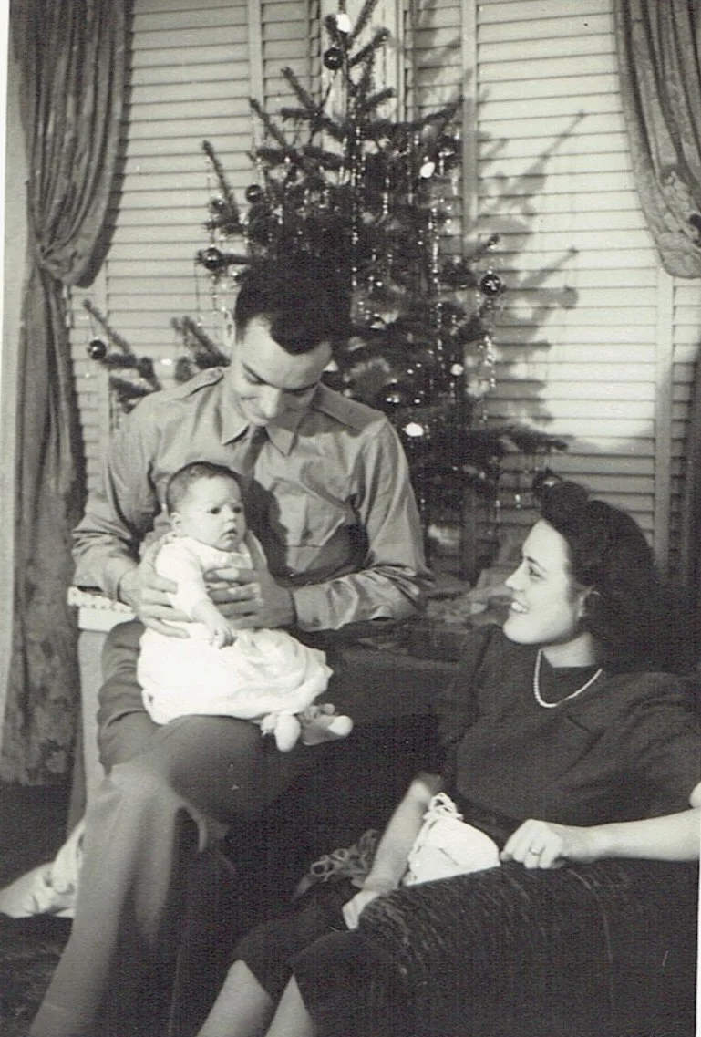 black and white picture of a judith mcdaniel's father sitting next to a christmas tree with baby judith in his lap, her mother is sitting on the floor smiling at them