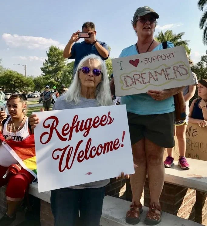Judith at a protest in Tucson, 2017 in response to Trump’s Muslim ban.judith mcdaniels with a sign that says "refugees welcome" judith is wearing sunglasses. a woman is next to her with a sign that says "i heart dreamers"