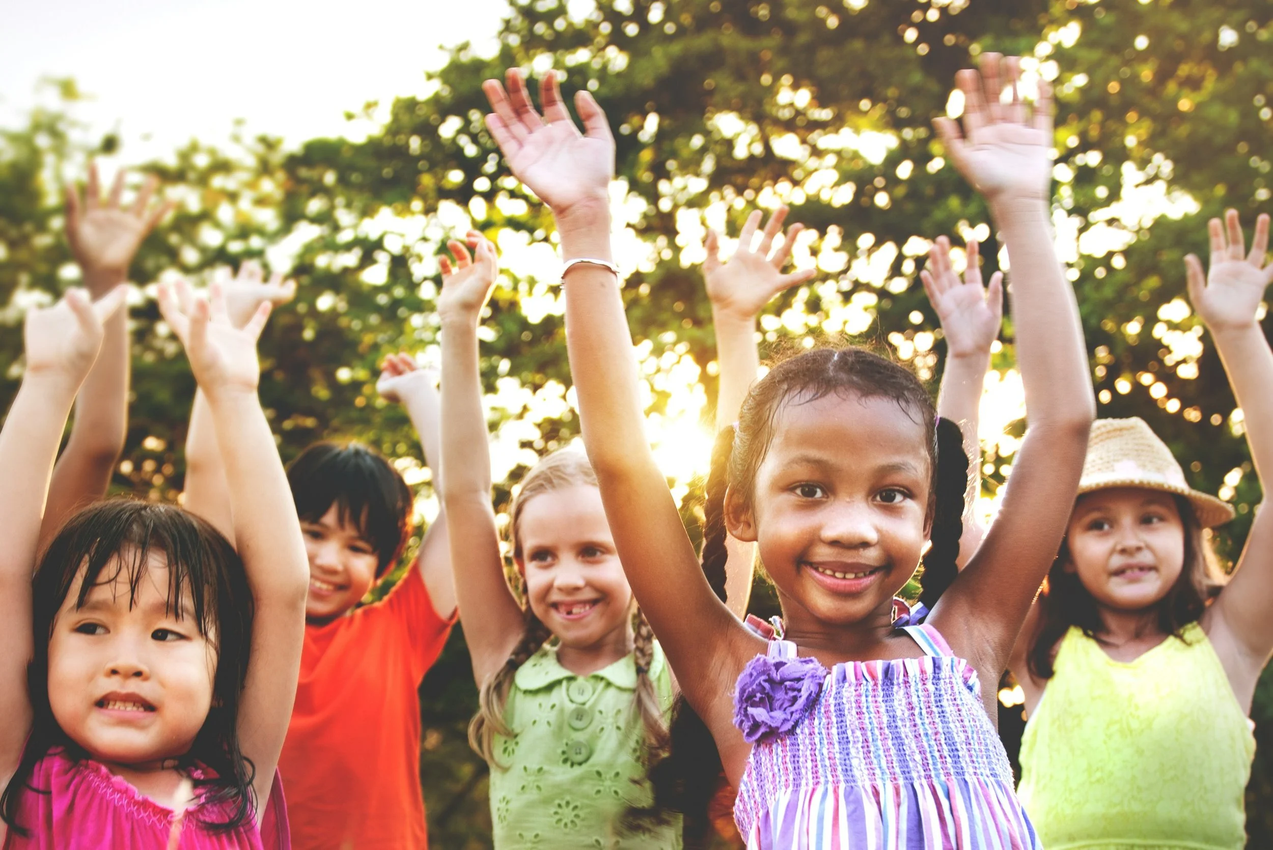 A group of children raising their hands outdoors in the sunlight, smiling and enjoying themselves in a park with trees in the background.