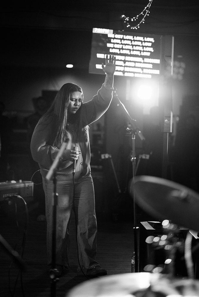 A woman with long hair and casual clothing singing into a microphone with her eyes closed, standing on stage in a dimly lit venue with a digital screen displaying song lyrics in the background.