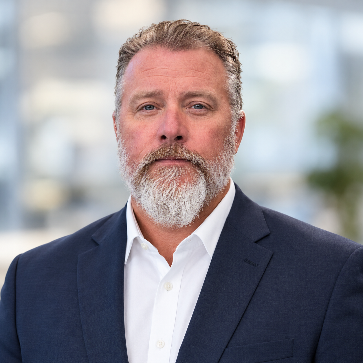 Close-up portrait of a middle-aged man with a beard wearing a navy blue suit and white shirt, standing indoors with a blurred background of windows and natural light.