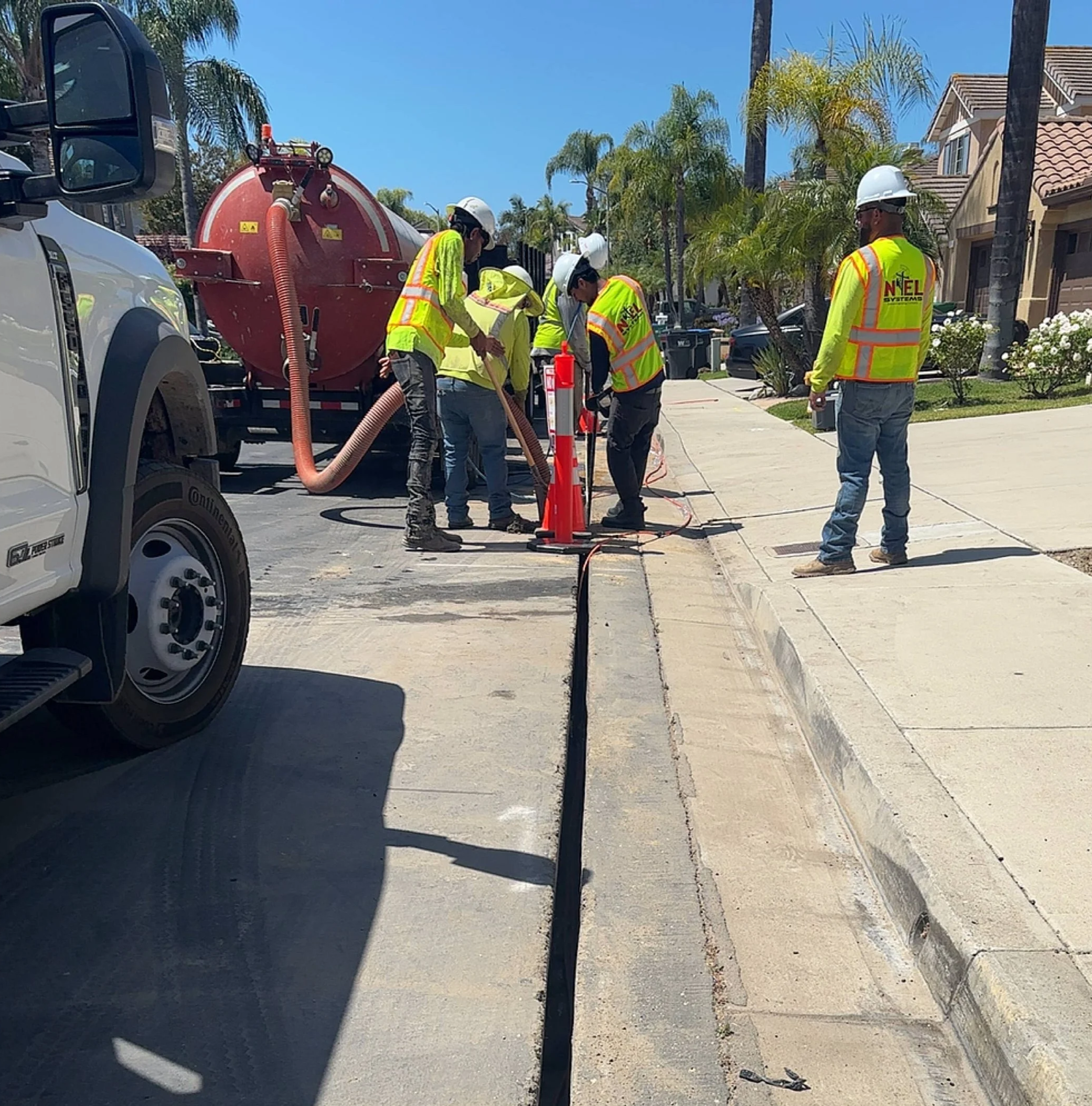 Construction workers wearing safety helmets and yellow reflective vests working on a sidewalk with construction equipment and orange cones on a sunny day, residential houses in the background, palm trees, and a clear blue sky.