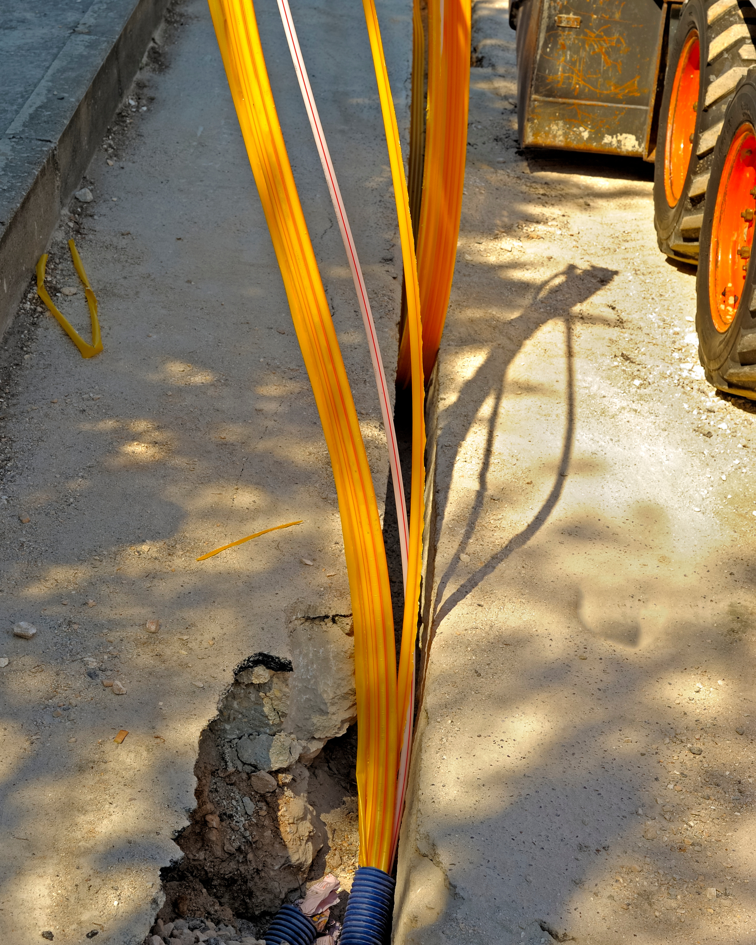 Yellow electrical conduit installed in a trench at a construction site, with black corrugated conduit connected at the bottom, and construction equipment with large tires in the background.