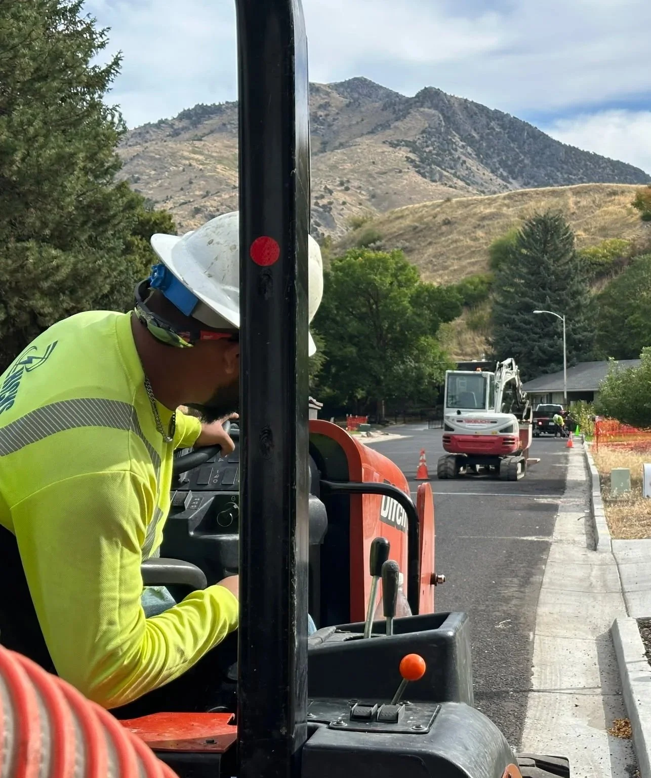 Worker in a neon yellow safety jacket, white hard hat with a blue device, operating a construction machine on a road with mountains and trees in the background.