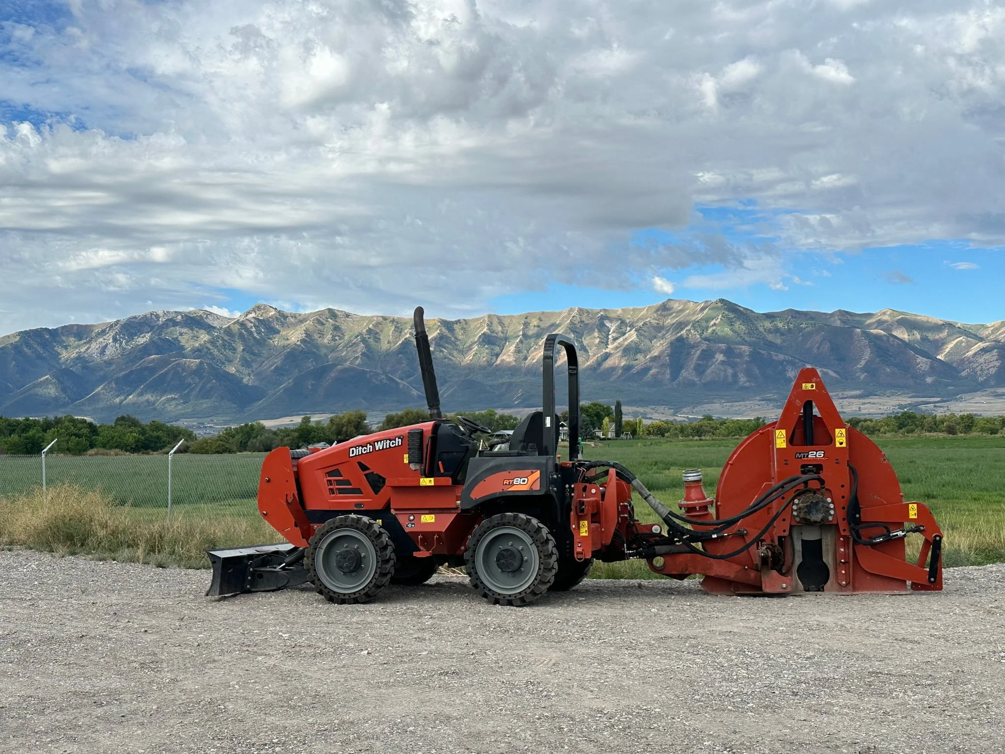 Red Ditch Witch turf cutter parked on gravel near a grassy field, with mountains and a partly cloudy sky in the background.