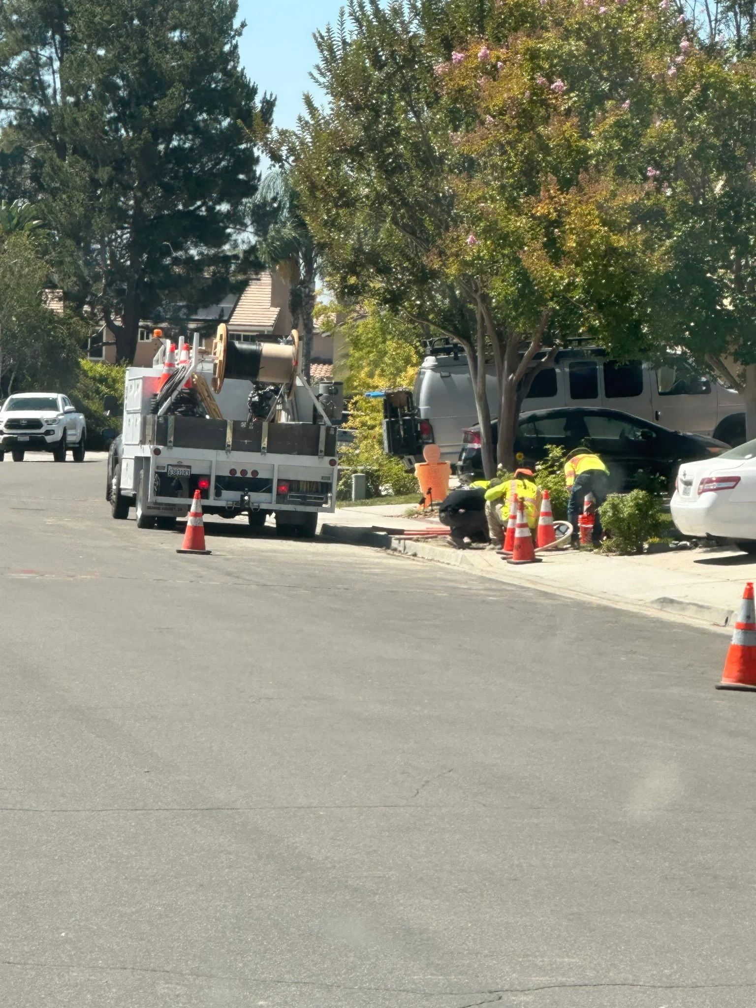Workers in safety vests and hard hats working on a sidewalk repair, with traffic cones and equipment, in a residential neighborhood with trees, parked cars, and houses in the background.