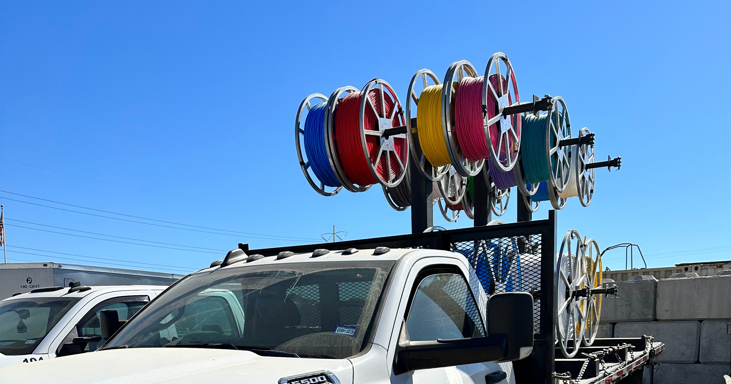 A flatbed truck with multiple spools of colored (blue, red, yellow, purple, green, white) filament or wire mounted on a rack at the back, set against a clear blue sky.