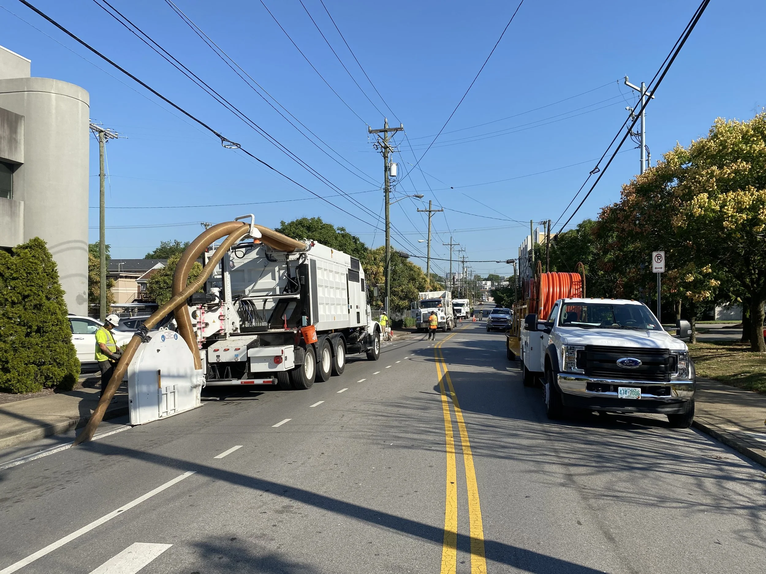 Street construction with workers and machinery, utility poles and power lines, trees, parked cars, and a clear blue sky.